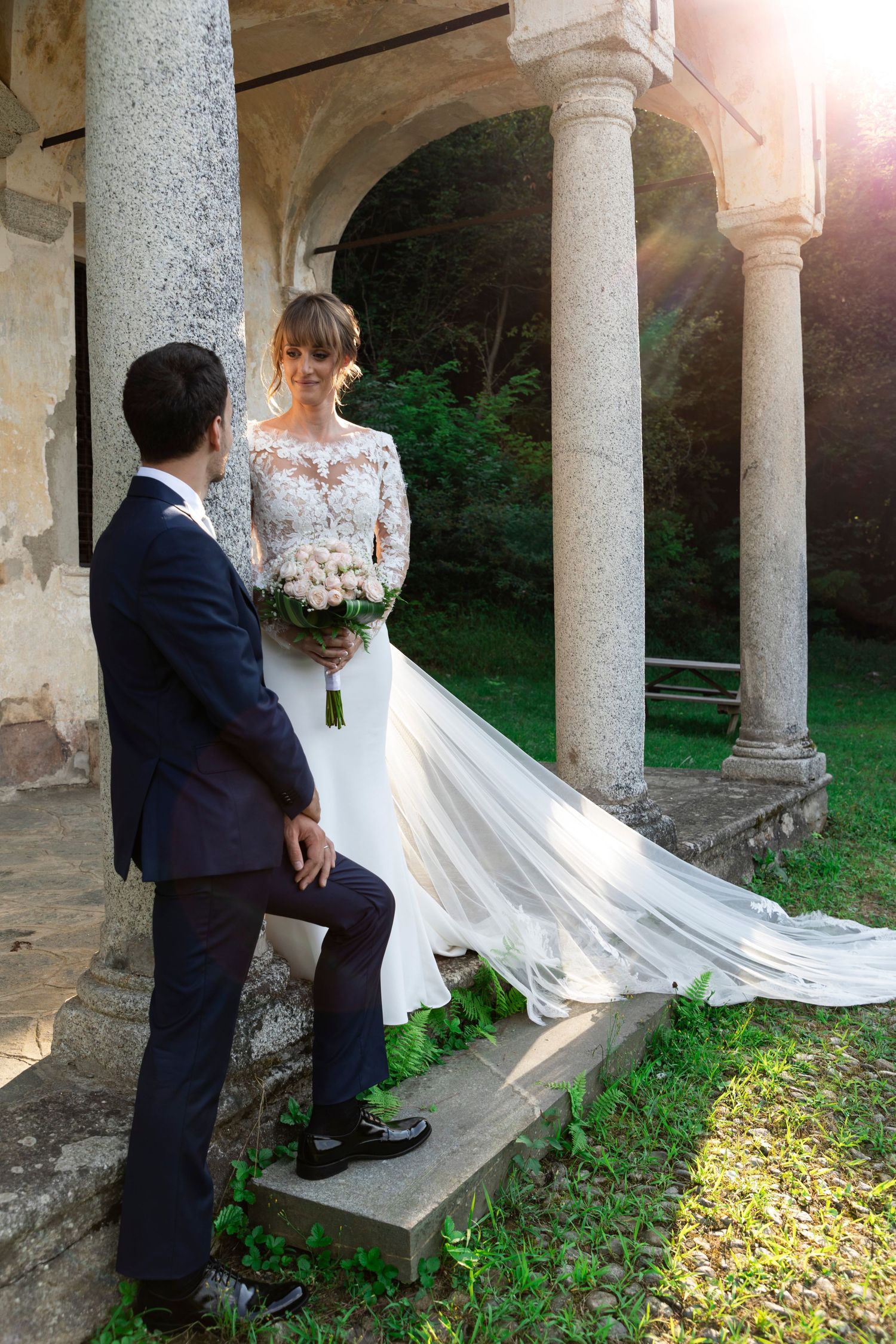 Bride in lace gown with flowing veil stands on stone steps with groom at sunlit colonnade.