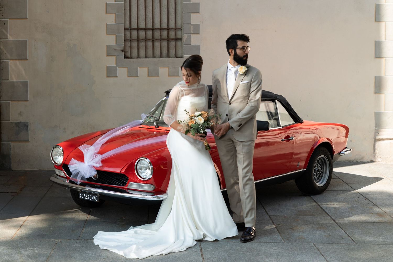 Stylish bride and bearded groom pose beside a classic red vintage convertible car on their wedding day.