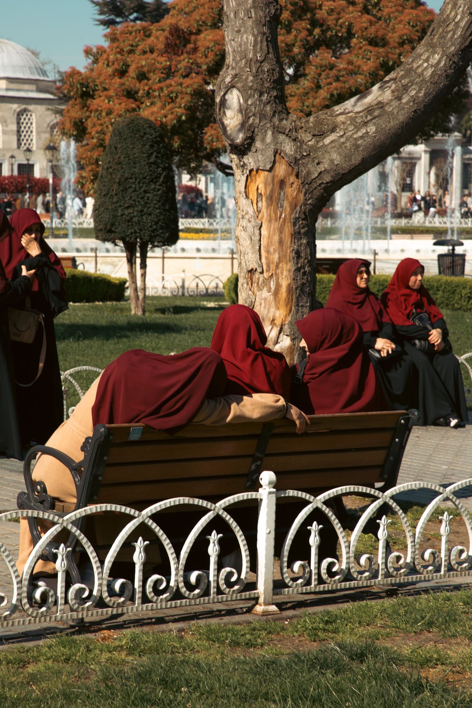 Women in dark red hijabs resting on an ornate white bench in a park near a mosque in Istanbul, Turkey.