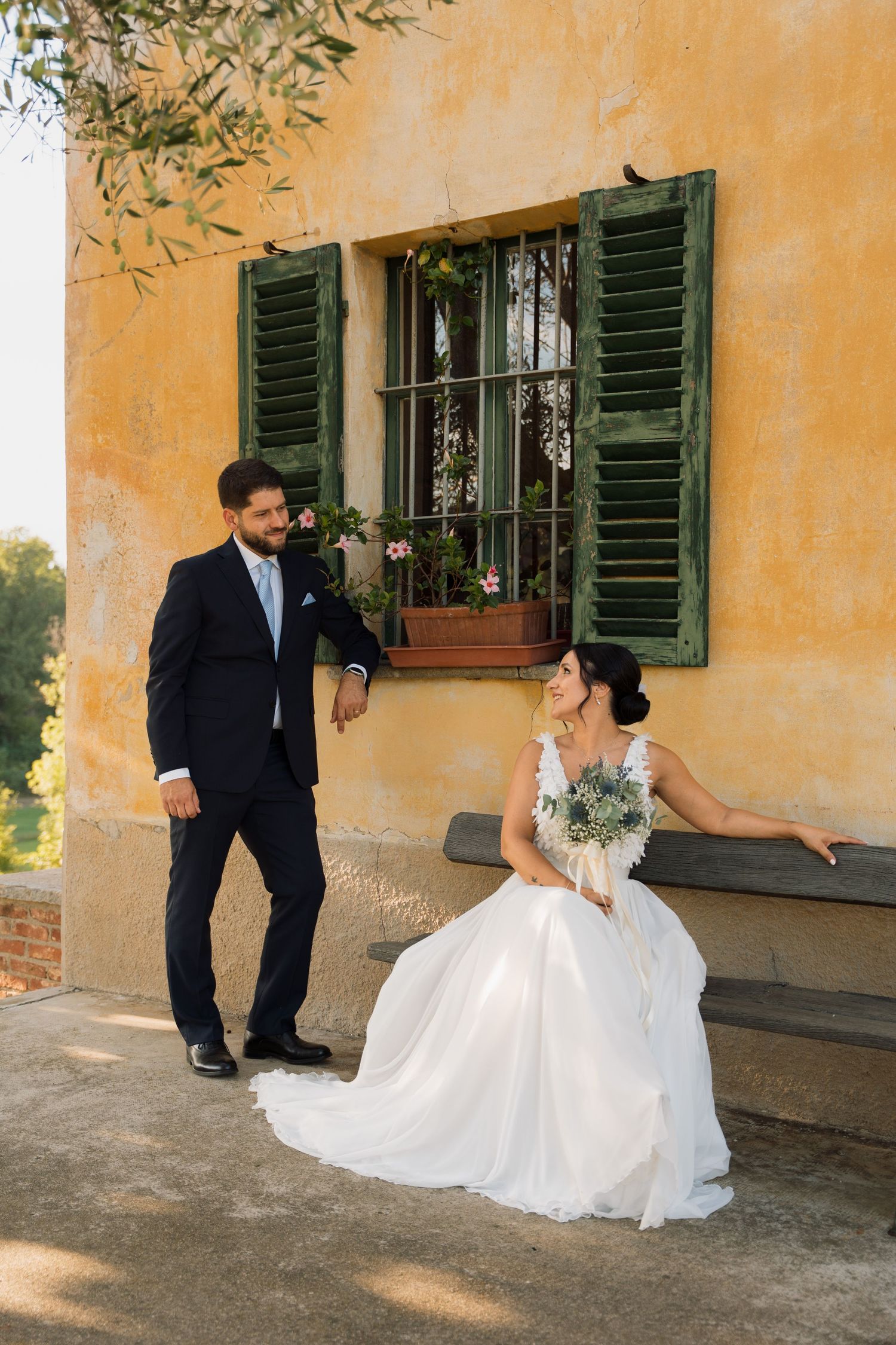 Bride in white gown sits on bench while groom in suit stands beside her at a rustic yellow villa.