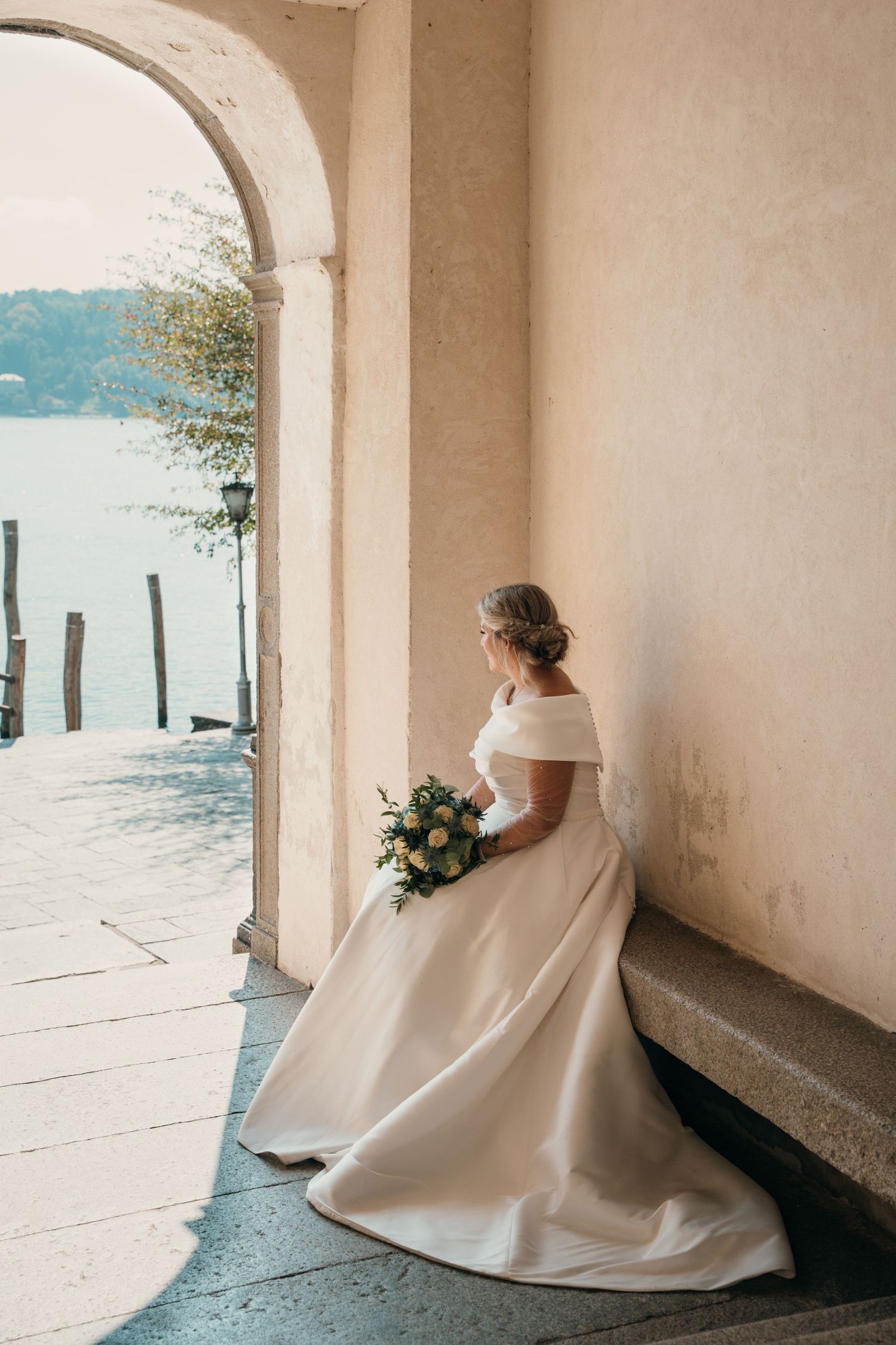 Elegant bride in off-shoulder gown holds bouquet while seated in arched colonnade overlooking serene waterfront.
