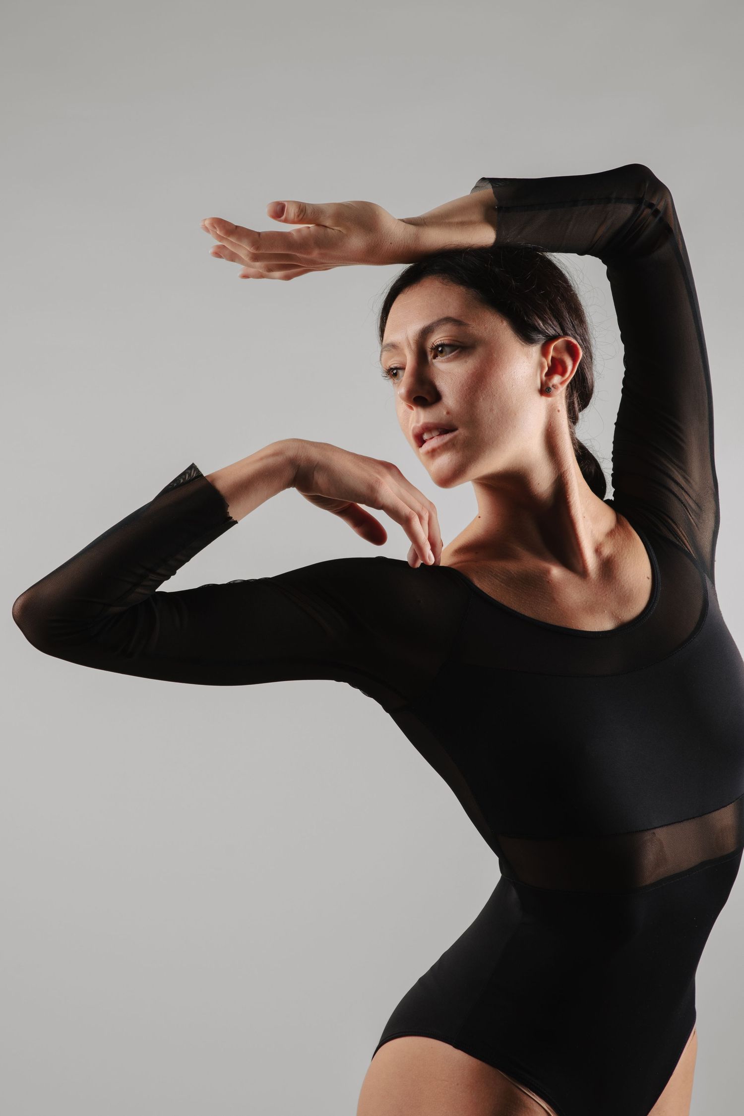 Female dancer in black long-sleeve leotard poses gracefully with arms raised in a studio setting.
