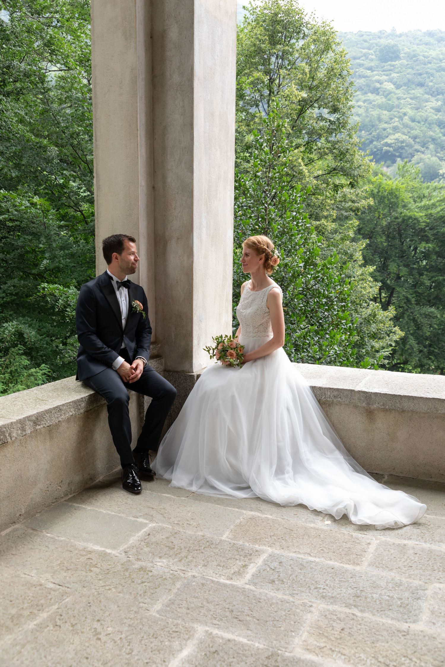 Bride and groom sit on stone balcony ledge surrounded by lush green forest, sharing a quiet moment.