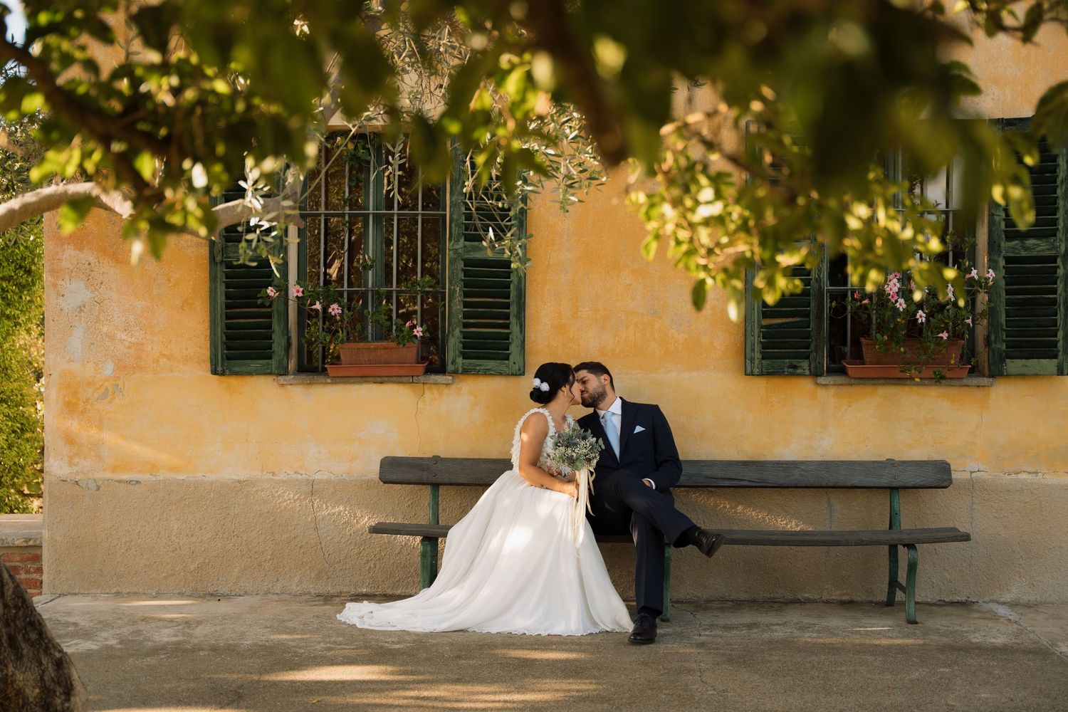 Bride and groom share a romantic kiss on a bench outside a rustic Italian villa surrounded by lush greenery.