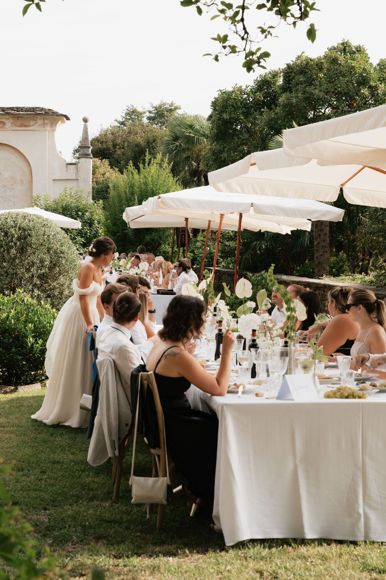 Elegant outdoor wedding reception with guests seated at long white-draped tables under cream parasols in a lush garden.