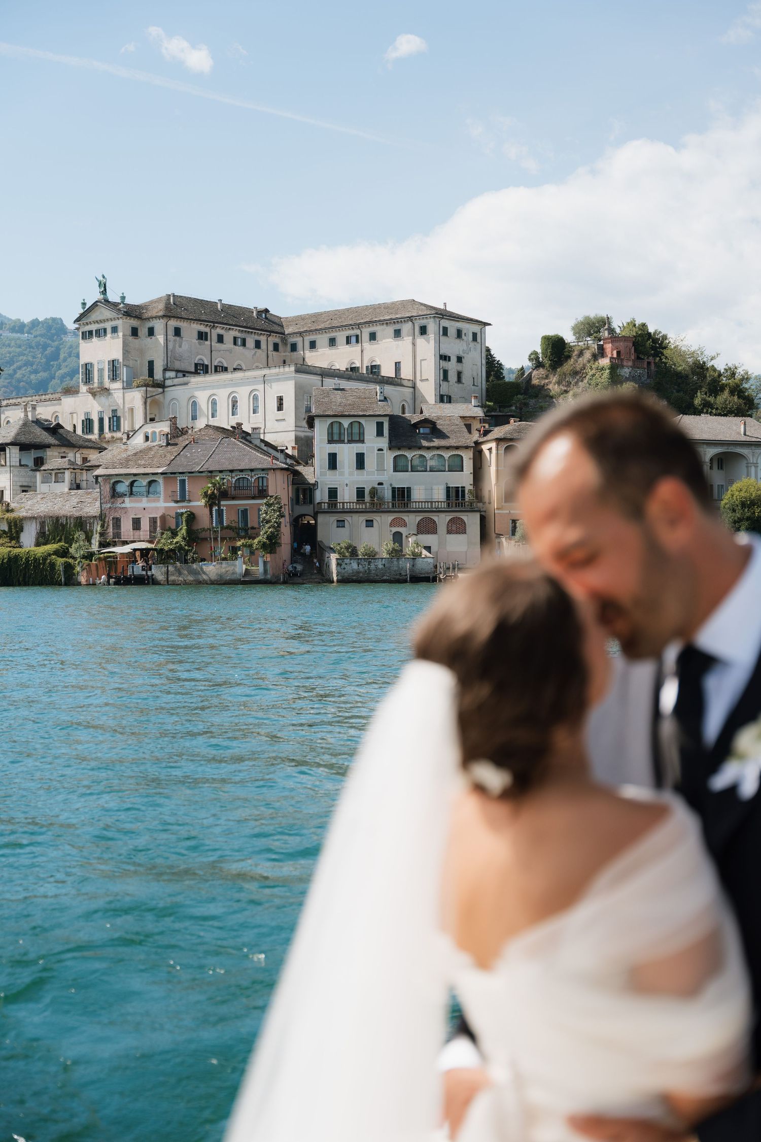 Bride and groom share a romantic moment on a boat with scenic Italian lakeside village buildings in the background.