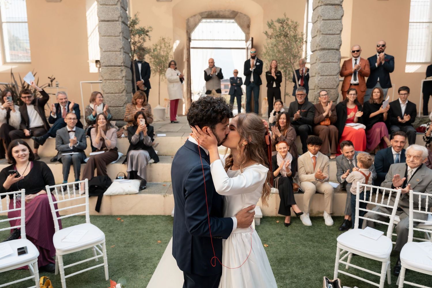 Newlyweds share first kiss as wedding guests applaud in a sunlit courtyard.