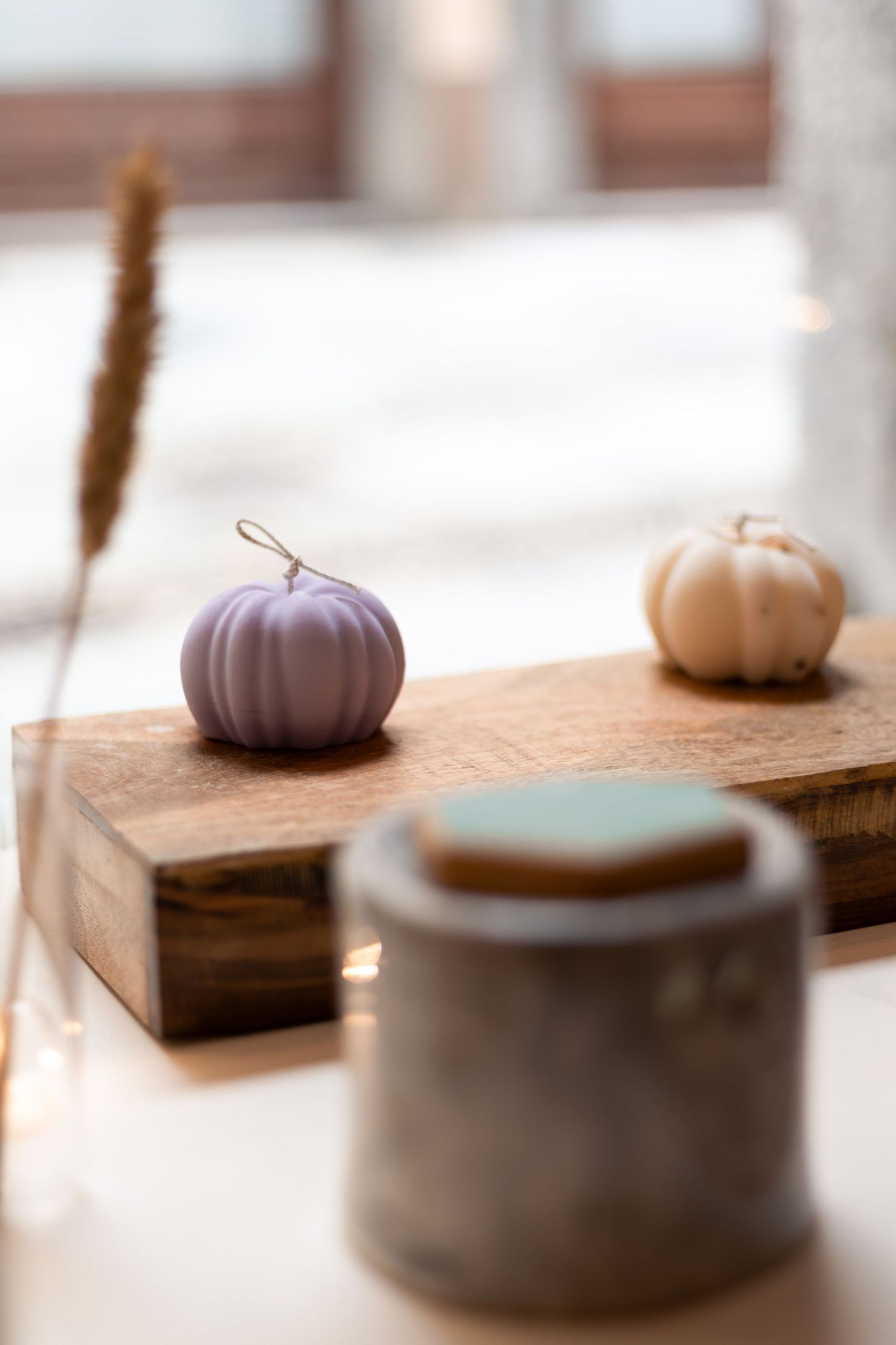 Two pumpkin-shaped candles in lavender and beige displayed on wooden board with bokeh lights.