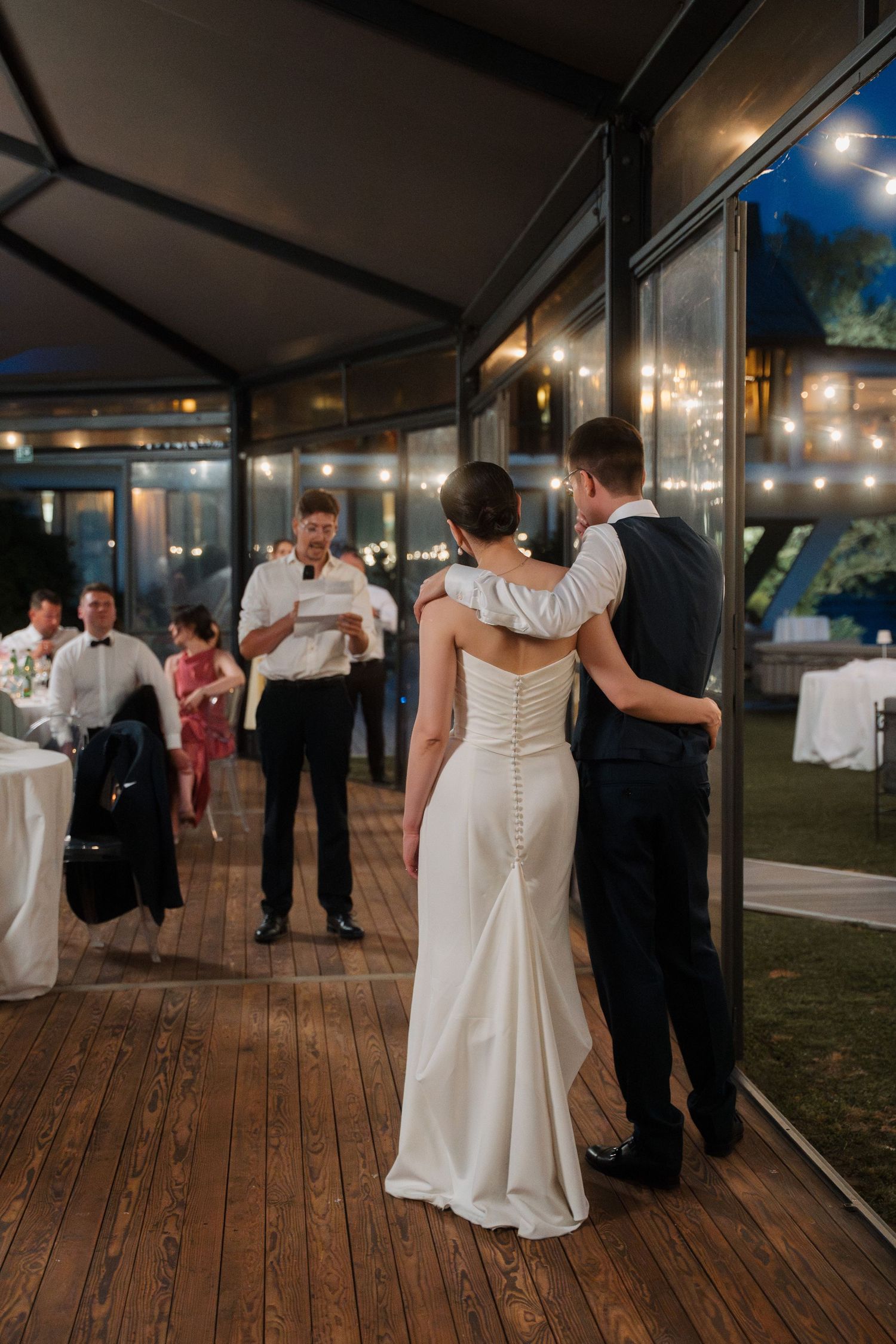 Bride and groom share their first dance on a wooden outdoor reception venue at night.