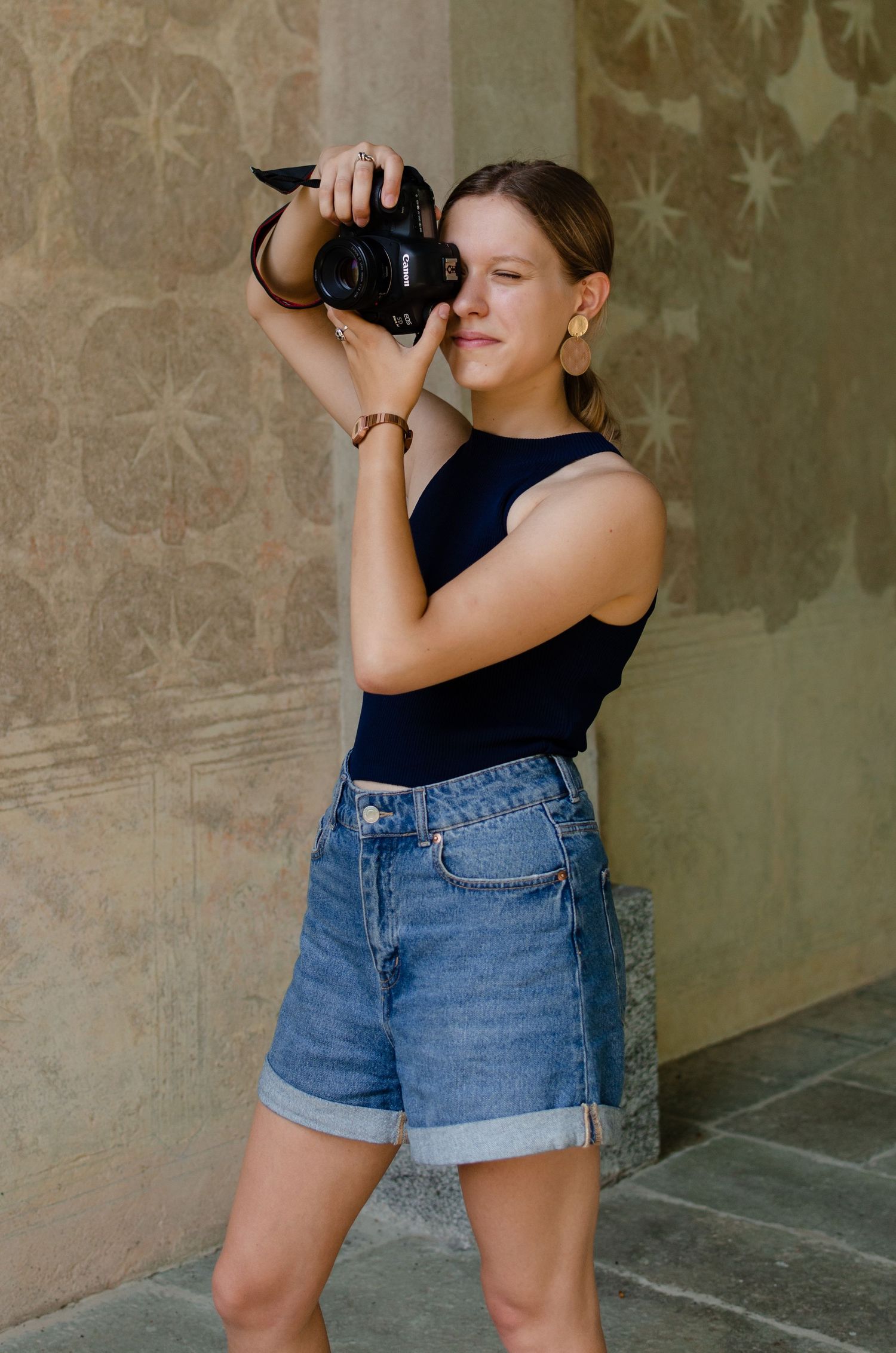 Young female photographer in navy top and denim shorts taking photos against decorative stone wall.