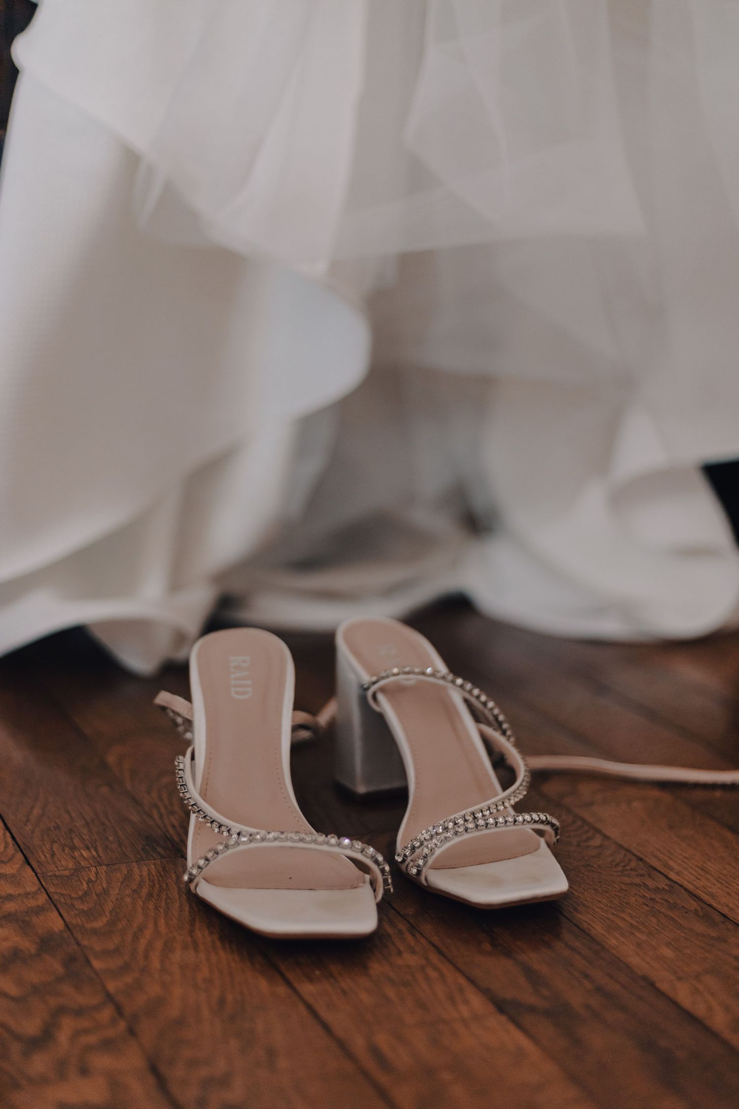 Elegant crystal-embellished bridal heels on hardwood floor beside flowing white wedding gown.