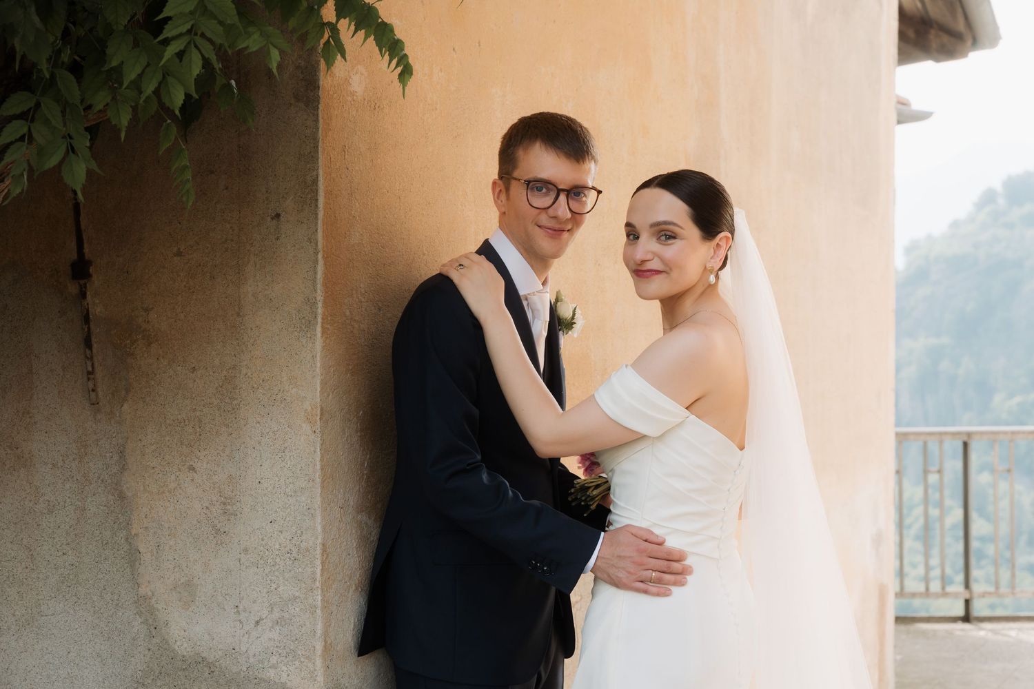 Bride and groom embrace near a rustic wall with scenic mountain views during their wedding photo session.