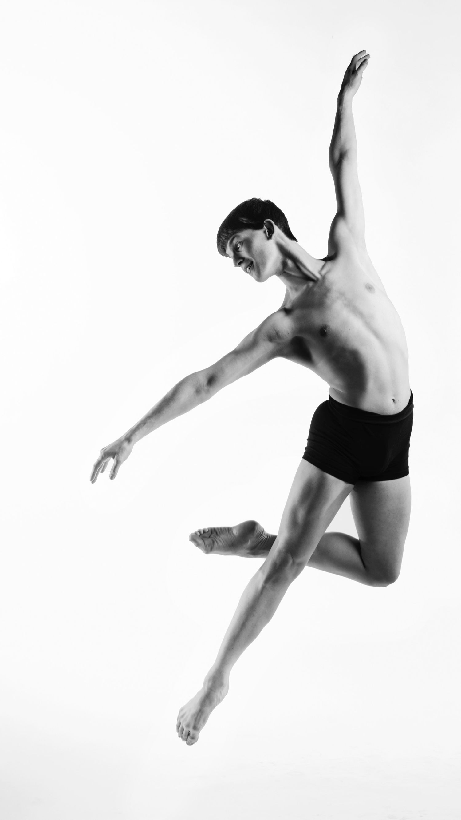 Athletic male dancer leaping dynamically in black shorts with arms outstretched against white background.