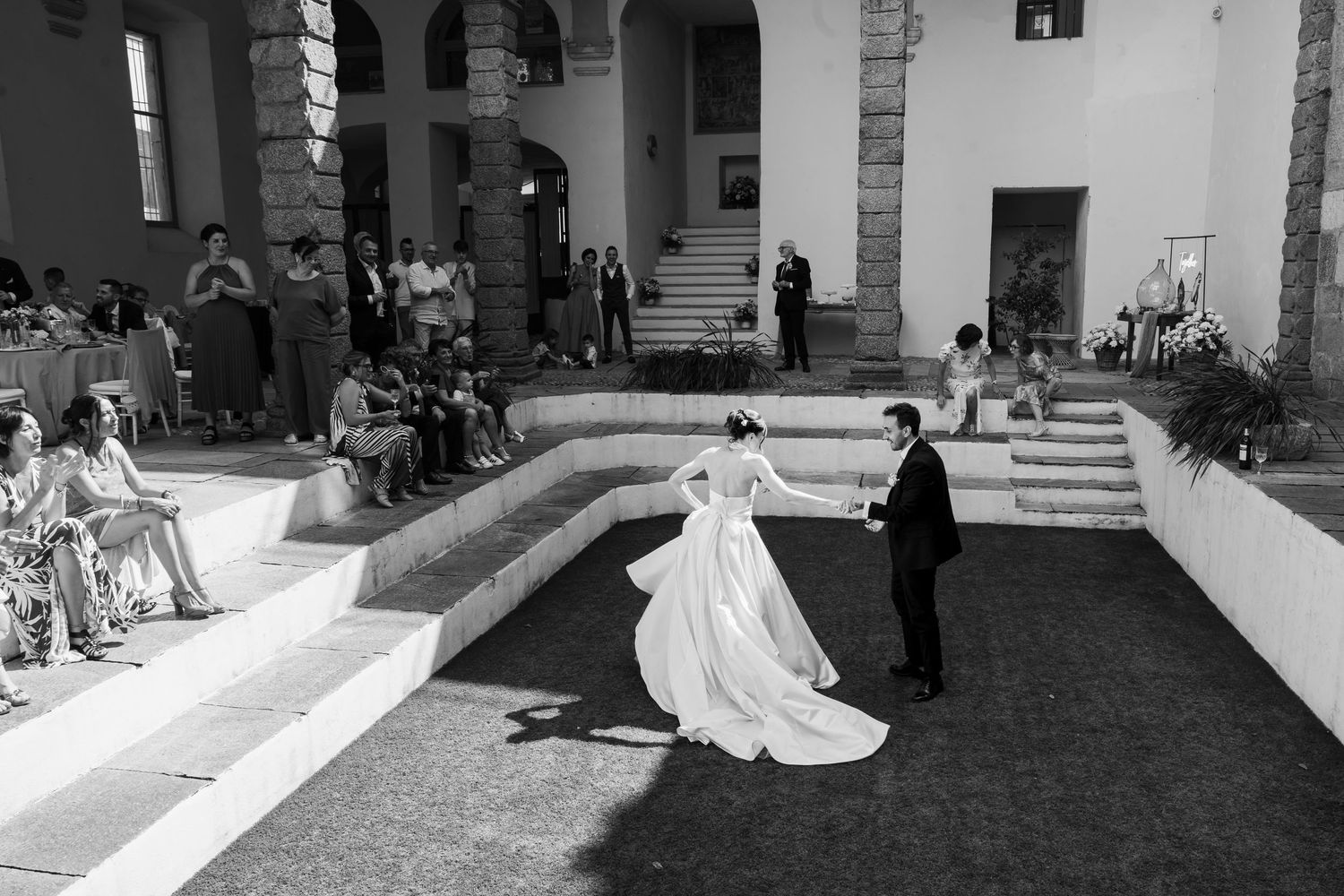 Bride in elegant gown and groom walk together in sunlit courtyard during black and white wedding ceremony.