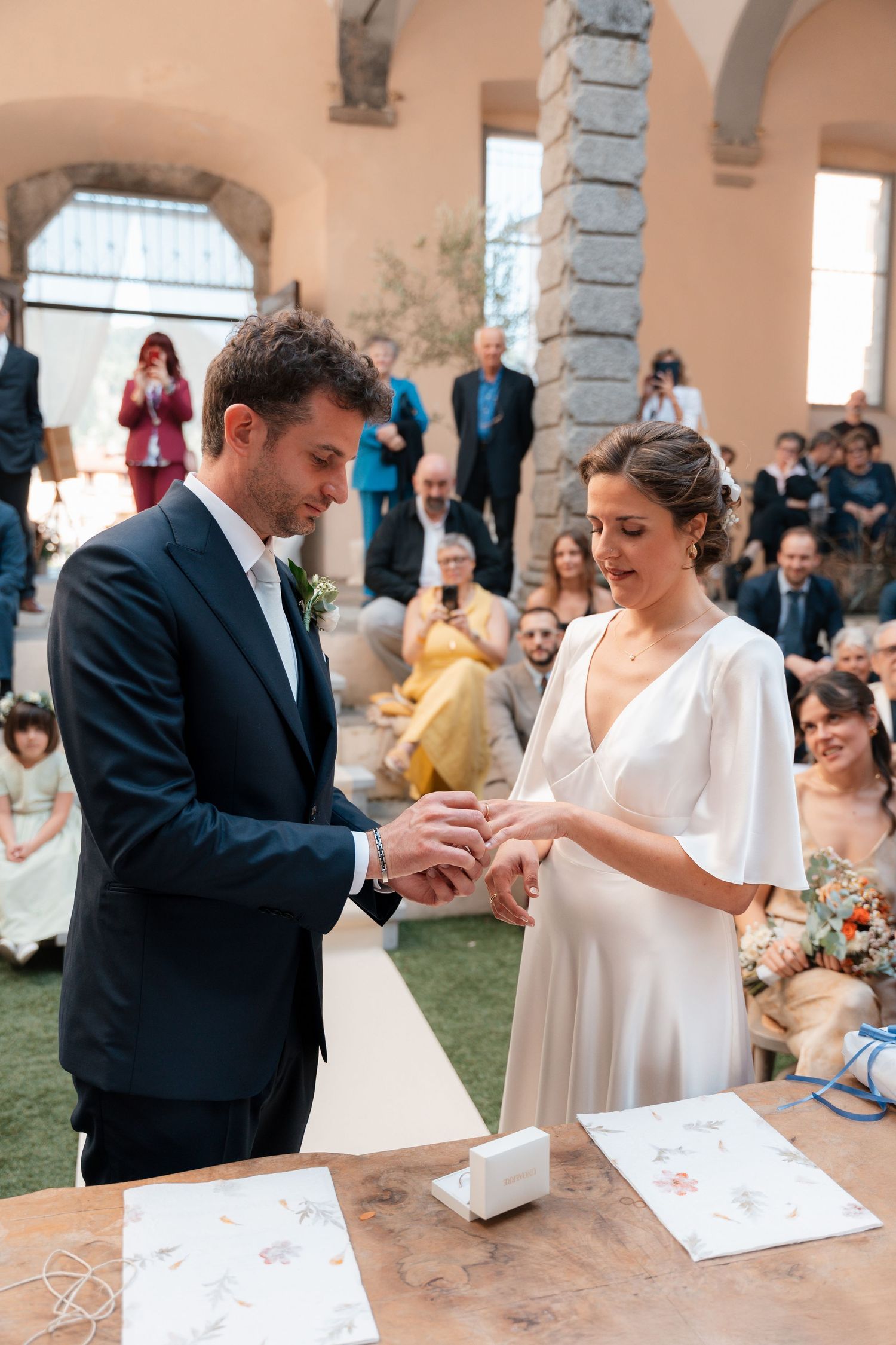 Groom places wedding ring on bride's finger at rustic wooden altar during outdoor ceremony.