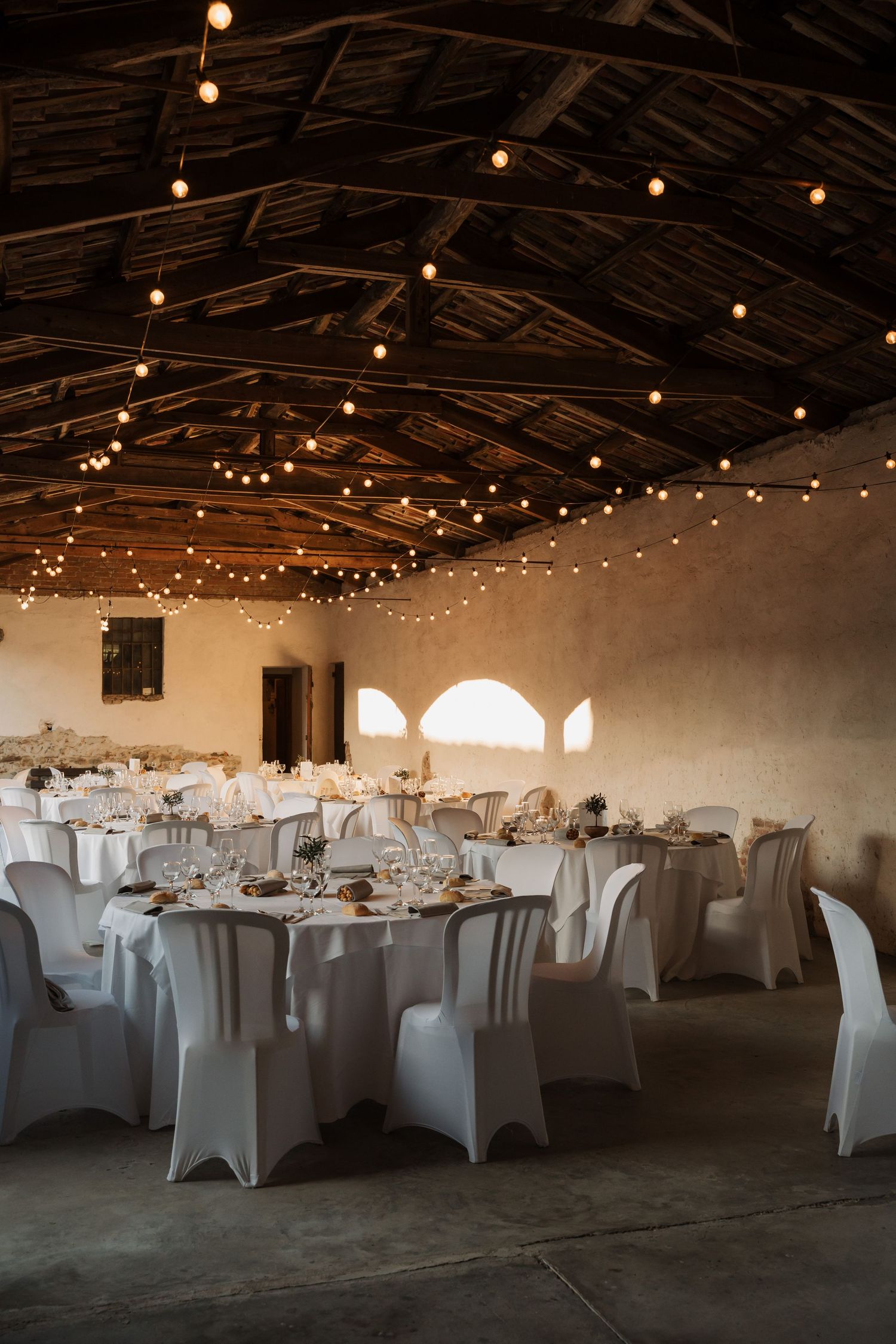 Elegant wedding reception hall with string lights, white draped chairs, and round tables set for dining.