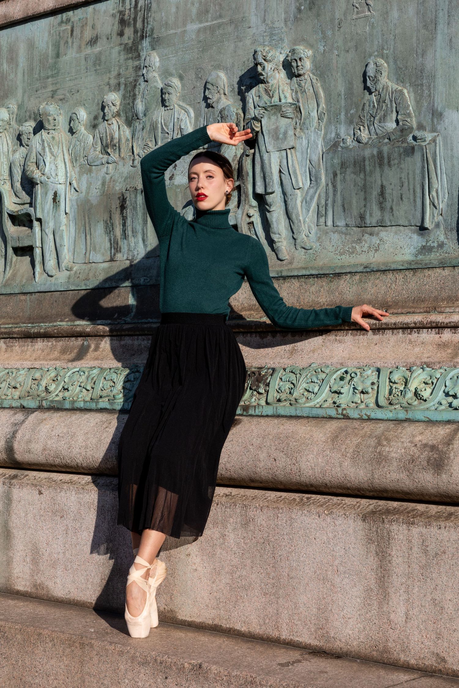 Ballerina in green turtleneck and black skirt posing on pointe before ornate bronze relief sculpture.