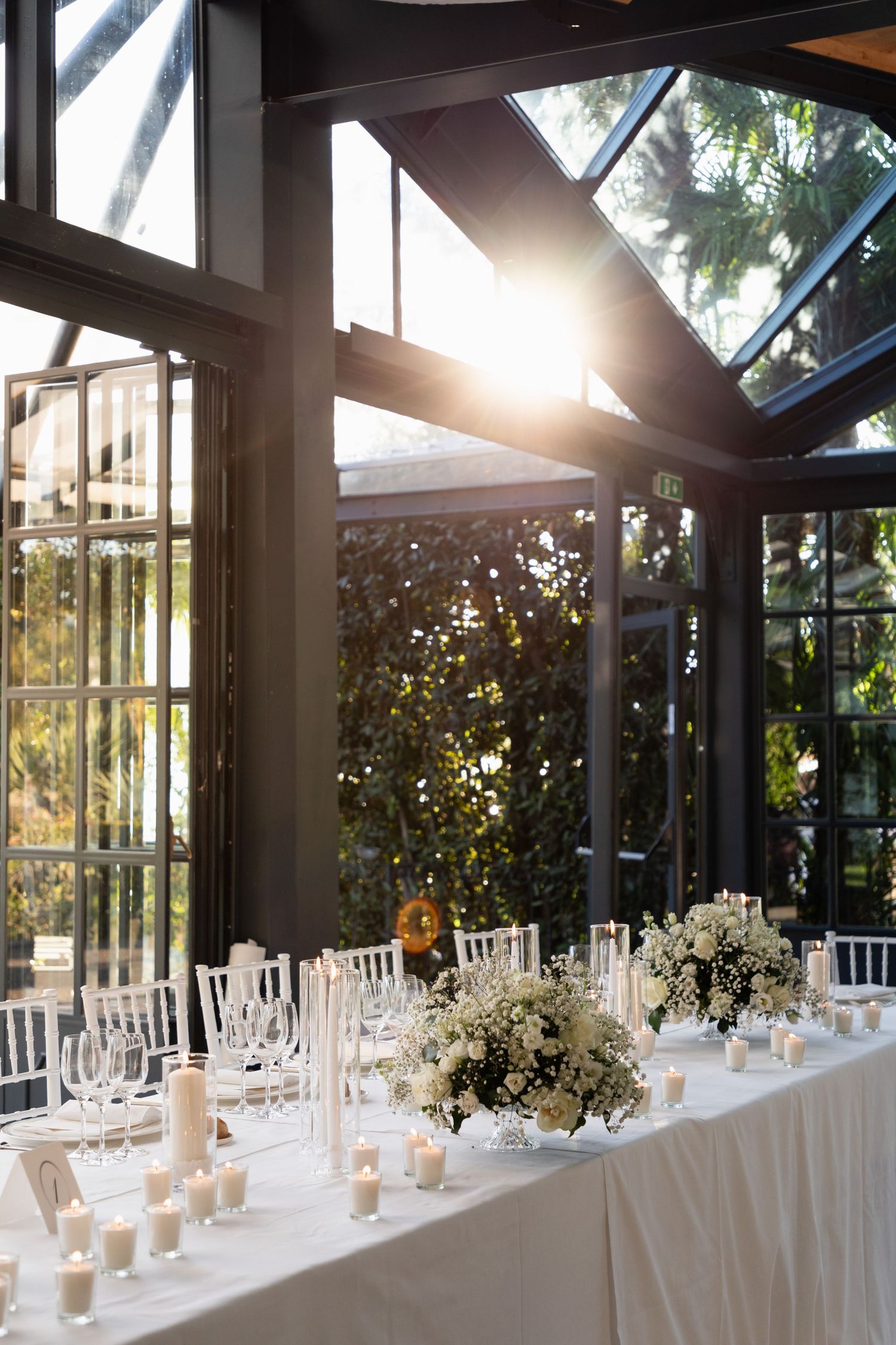 Elegant wedding reception table with white floral arrangements, candles, and crystal glassware in a sunlit greenhouse venue.