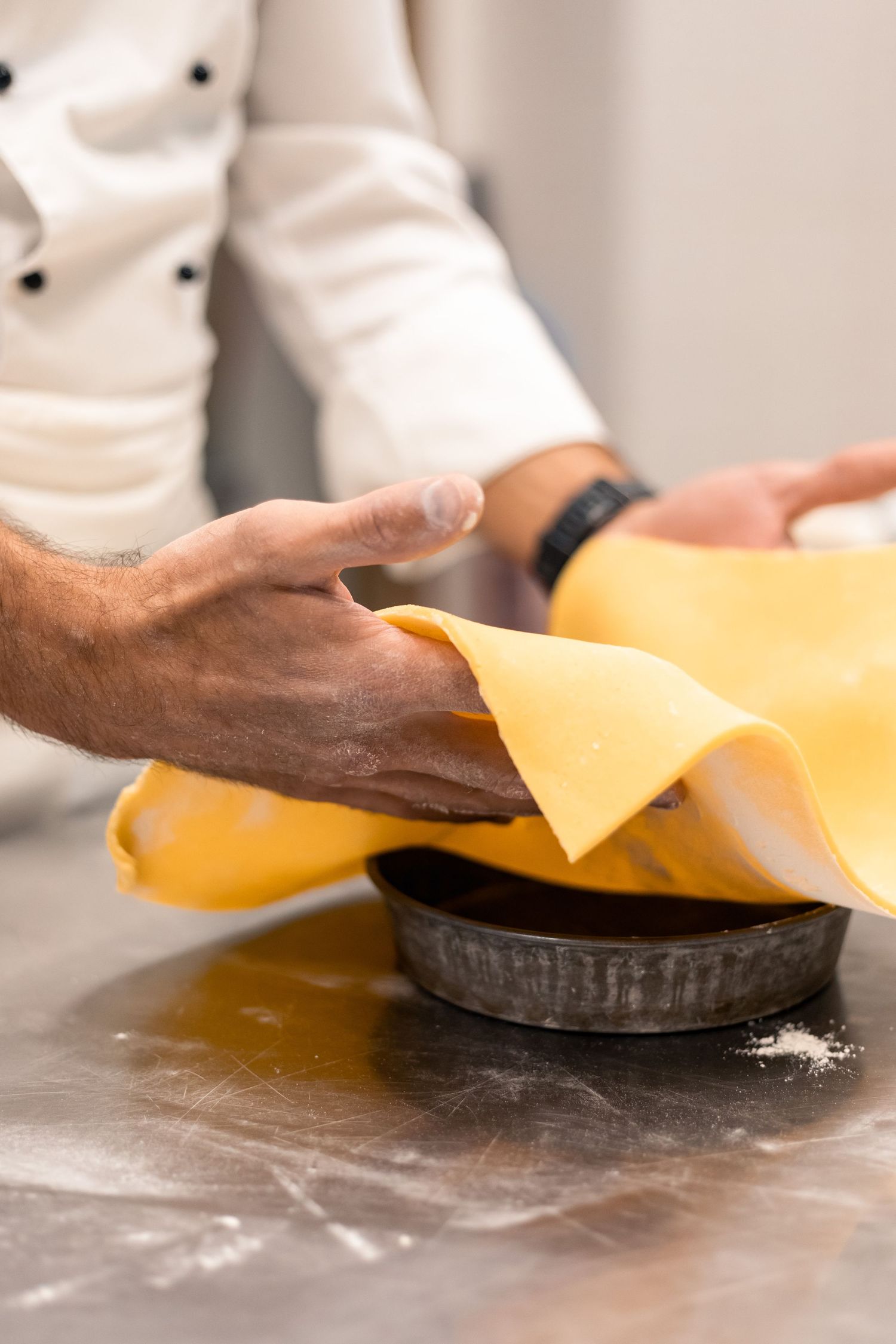 Chef placing thin yellow pasta dough sheet into dark baking pan on flour-dusted metal surface.