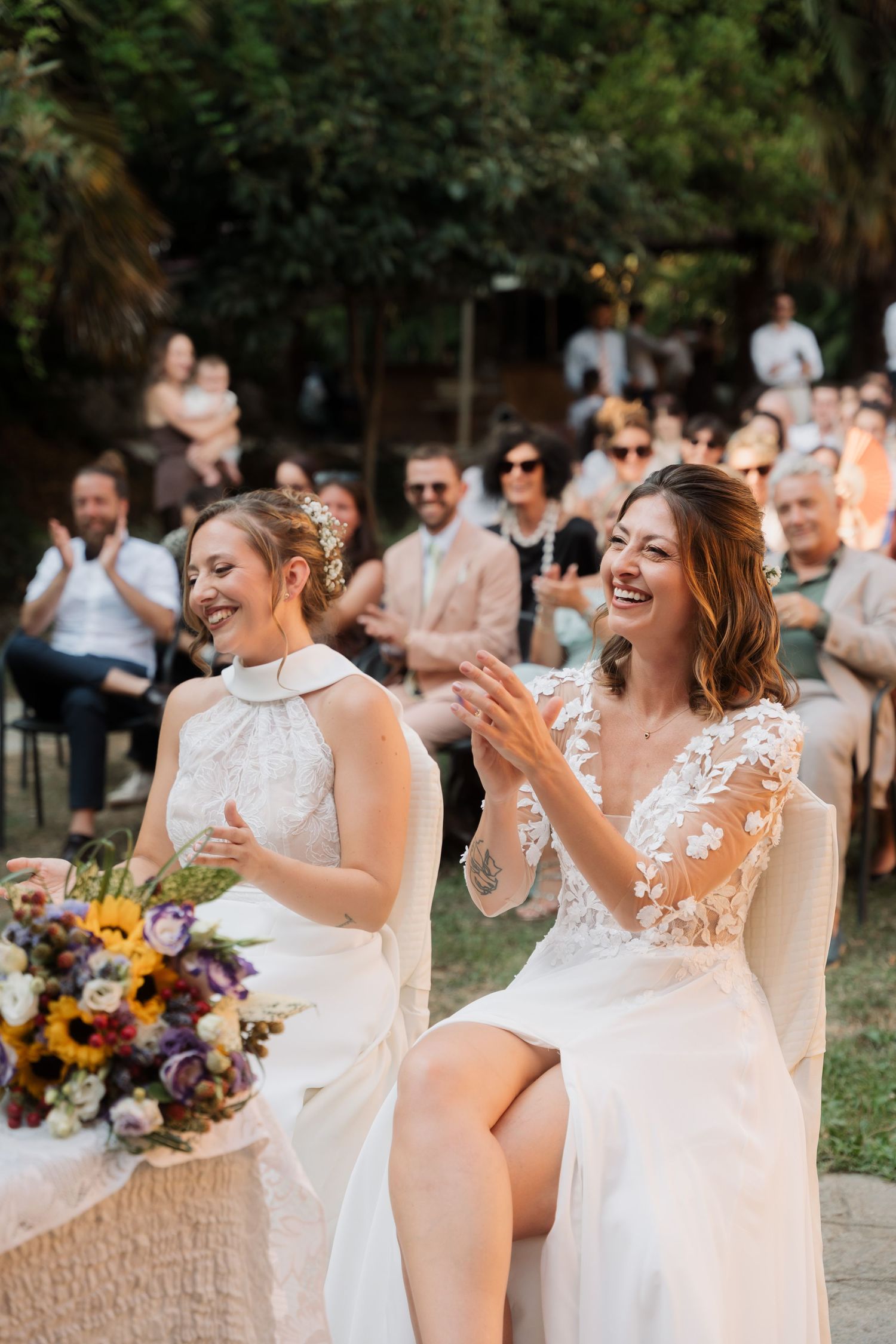 Two brides in white gowns laugh joyfully at their outdoor wedding ceremony, surrounded by clapping guests.
