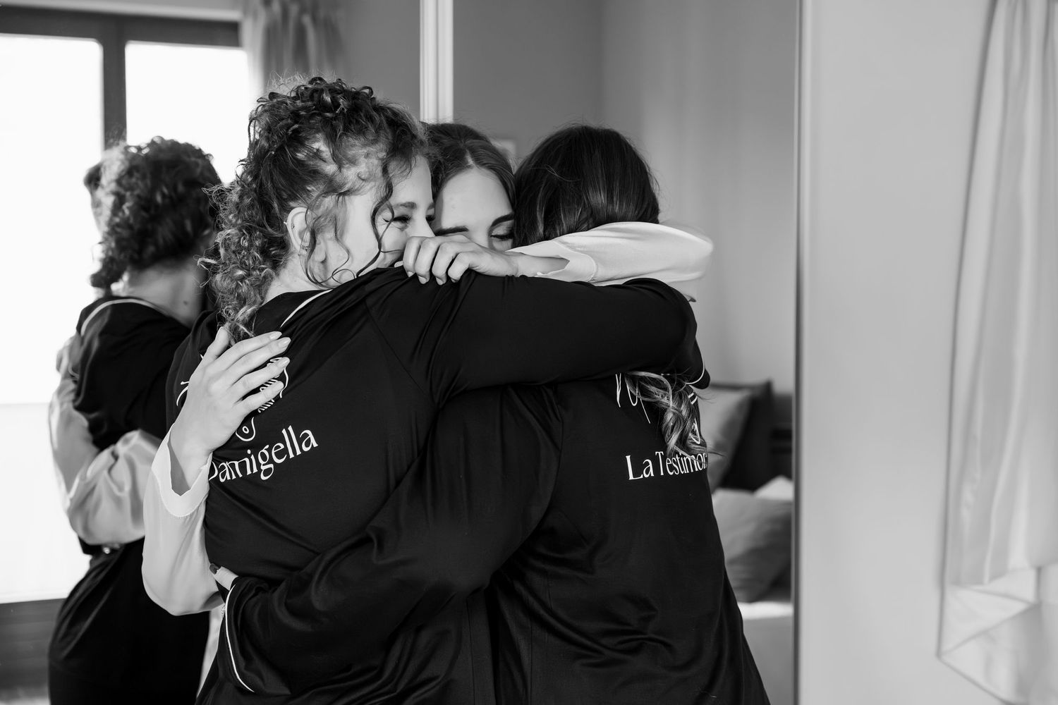 Emotional black and white photo of bridesmaids in matching robes sharing heartfelt group hug.