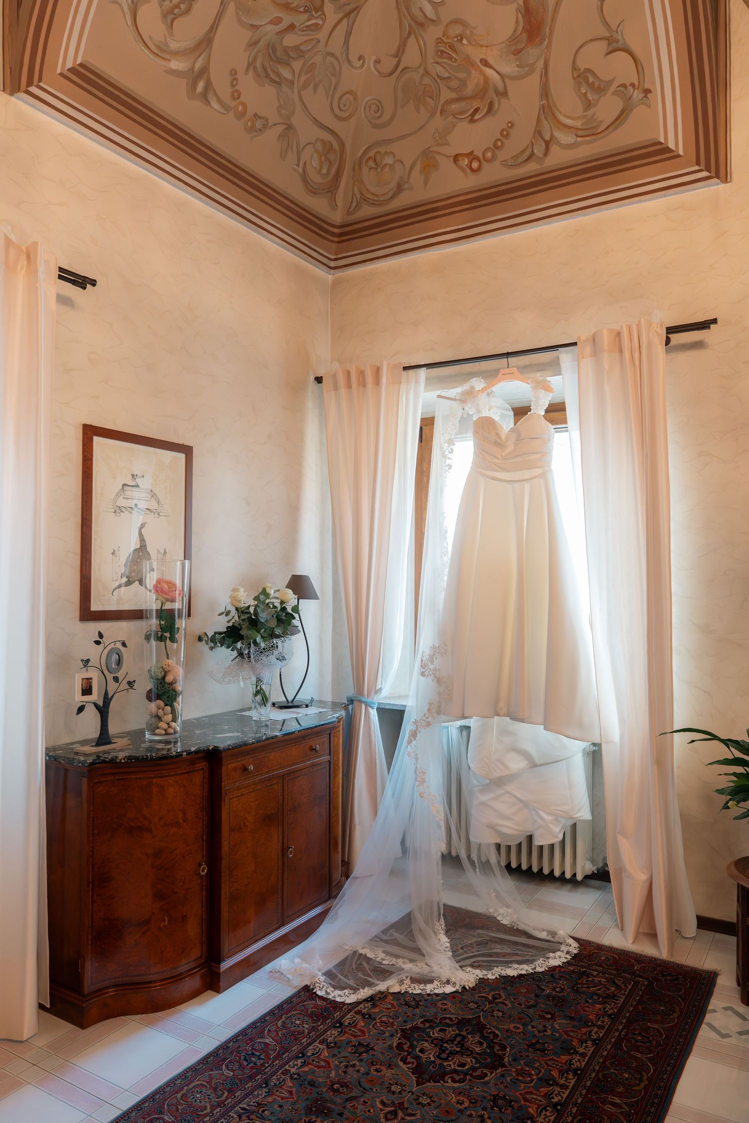 Elegant wedding dress hanging by window in ornate Italian villa room with frescoed ceiling.