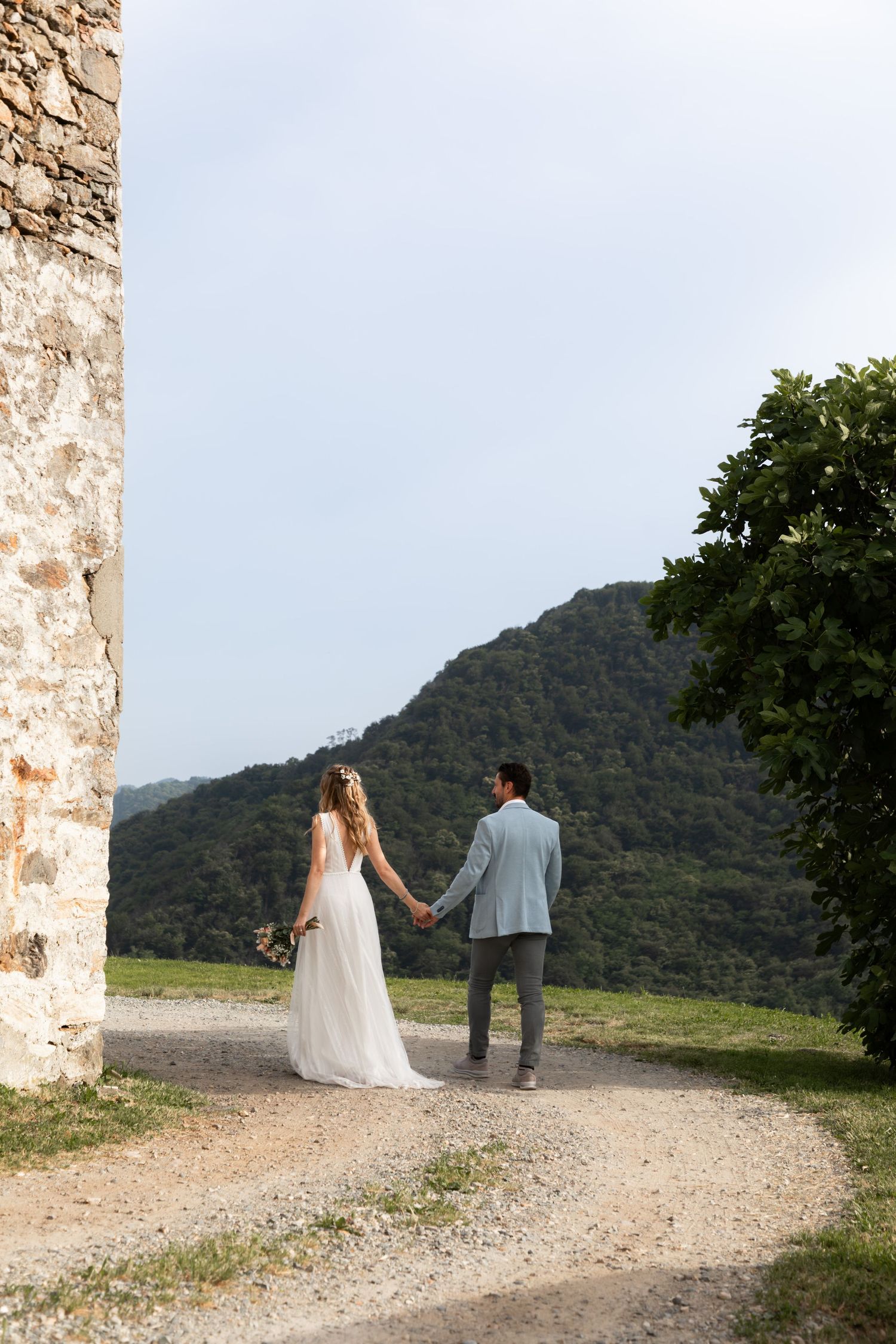 Bride and groom holding hands walking on gravel path beside stone ruins with green mountain landscape.