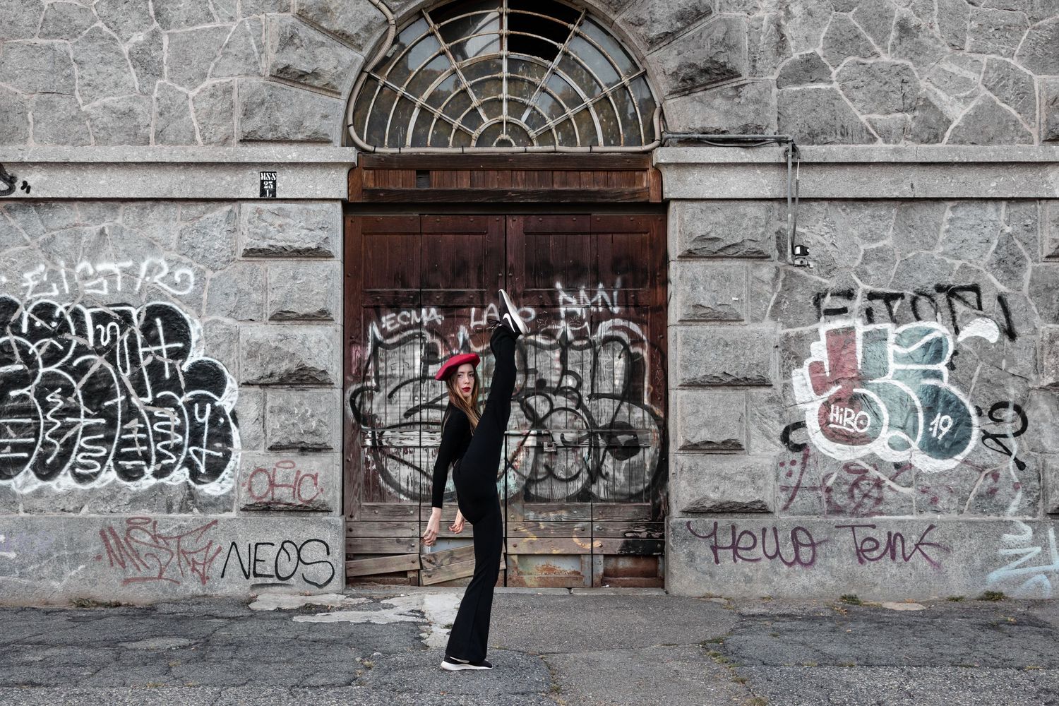 Person in black outfit with red beret dancing before graffiti-covered white building with wooden door.