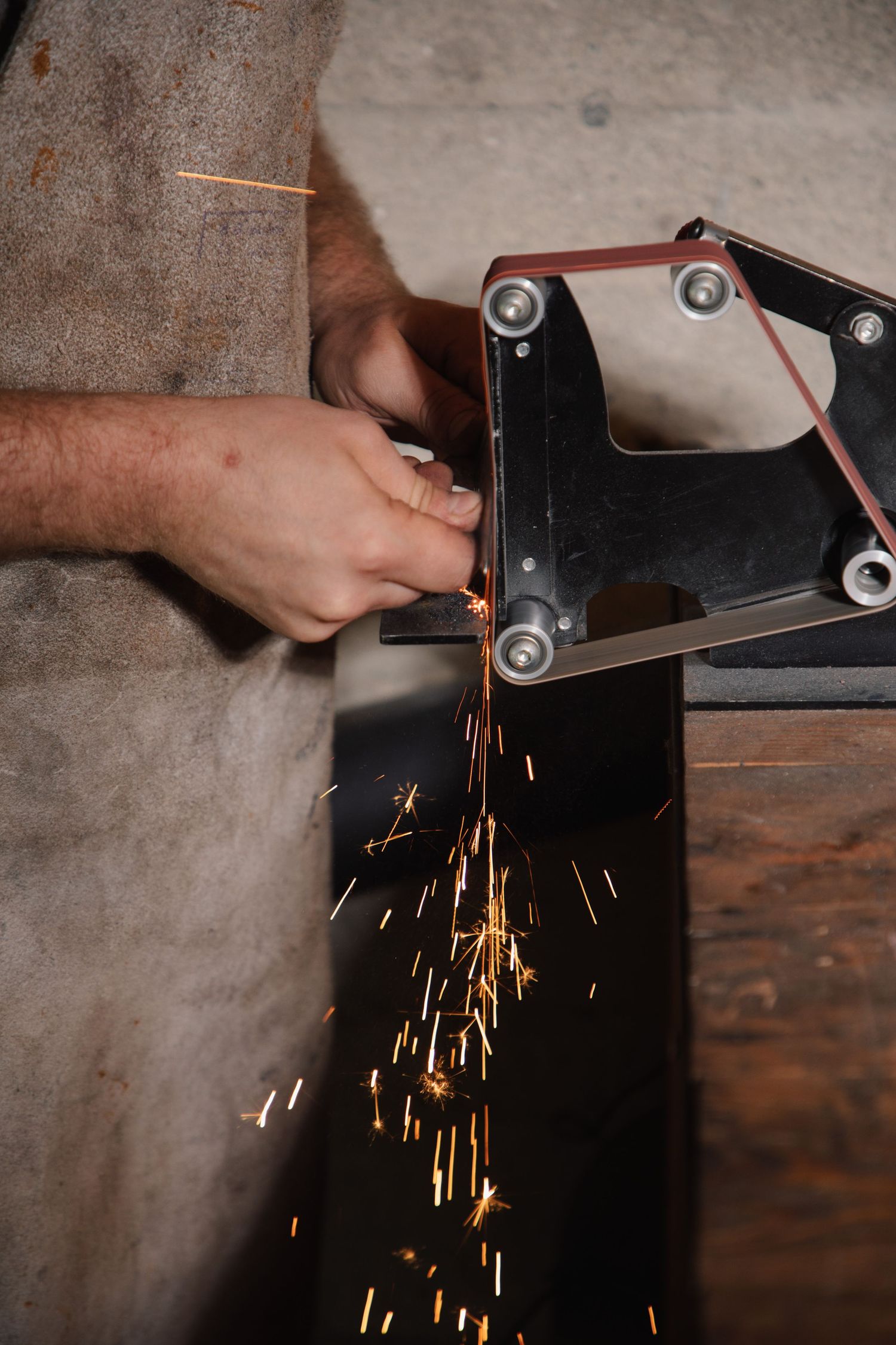 Craftsman sharpening metal blade on belt grinder with bright orange sparks flying in workshop.