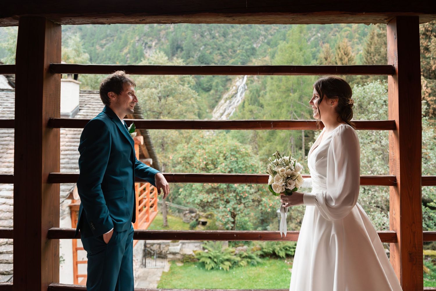 Bride and groom sharing a romantic moment by a wooden window overlooking a lush green mountain landscape.
