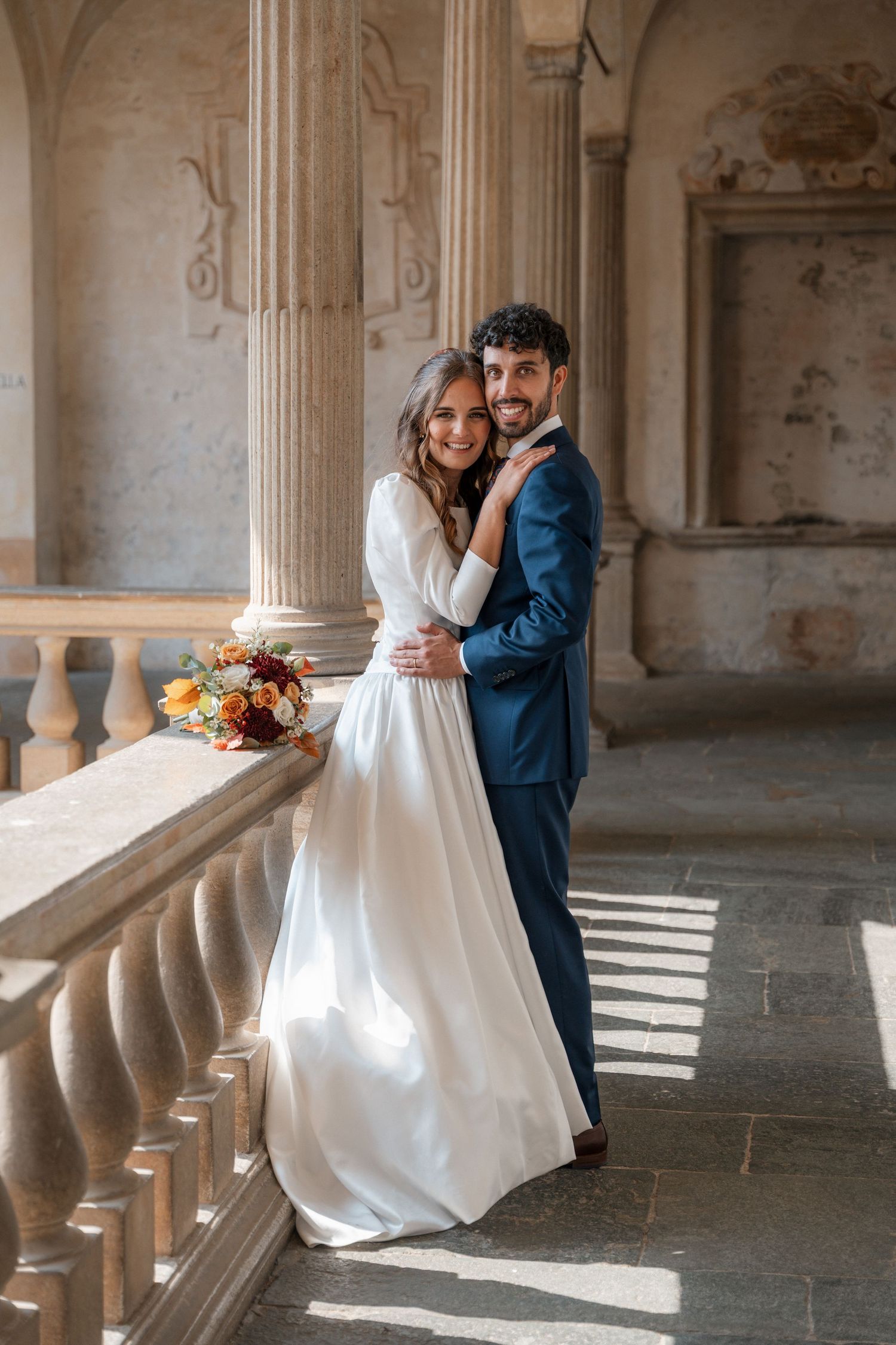 Smiling bride and groom embrace on sunlit classical colonnade balcony with autumn floral bouquet.