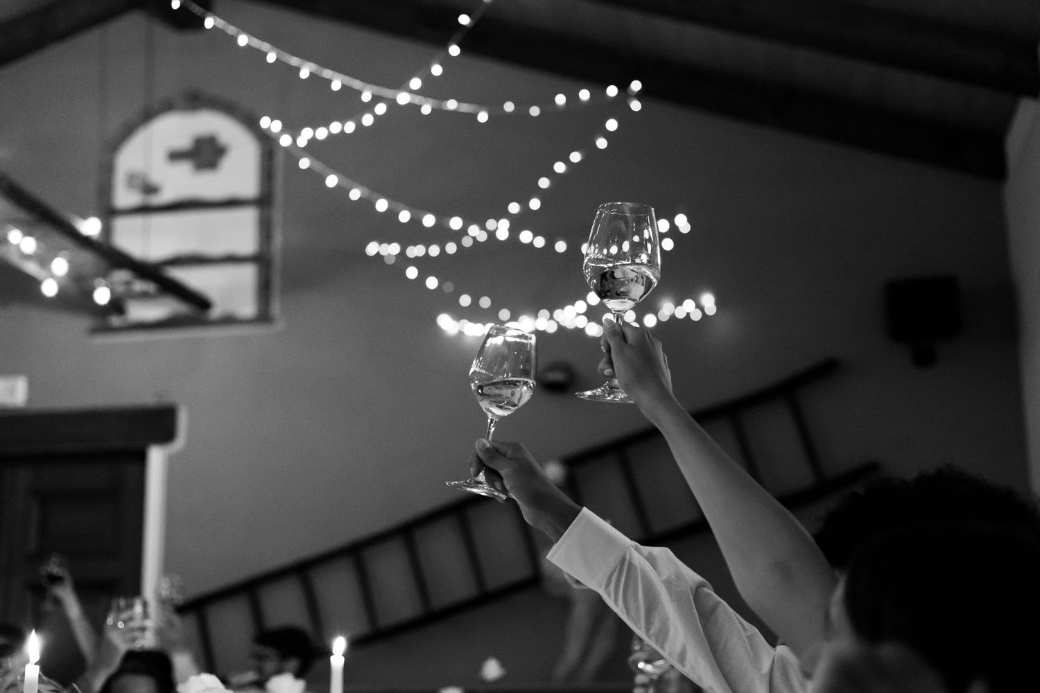 Guests raise champagne glasses in a celebratory toast under string lights at a wedding reception in black and white.
