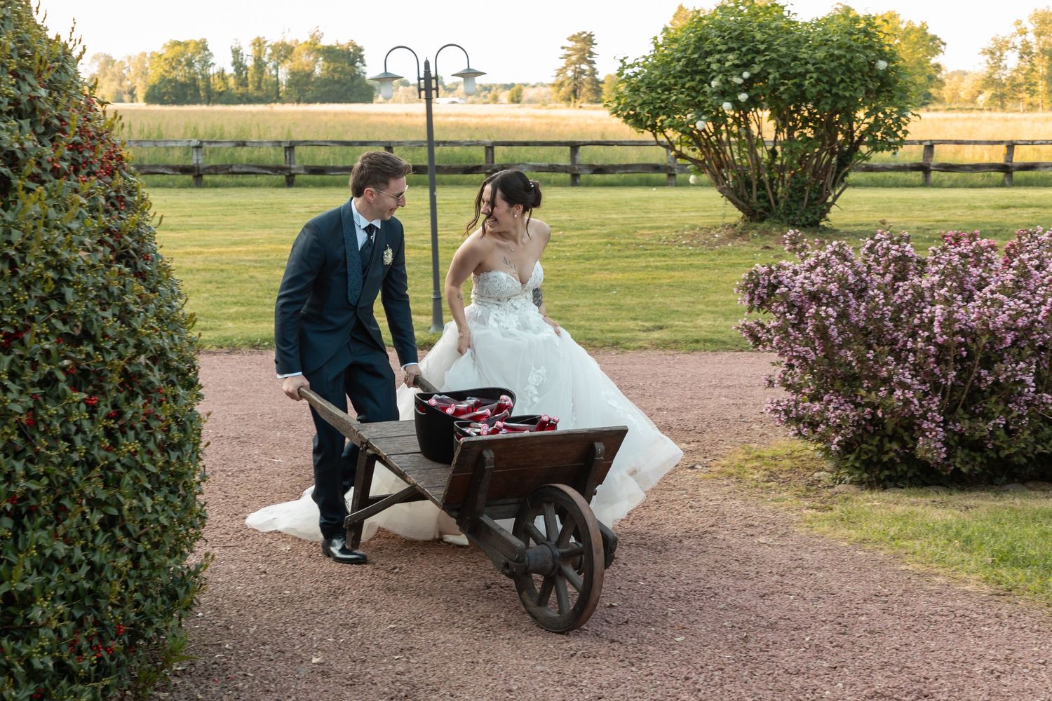 Groom playfully pushing bride in wooden wheelbarrow through elegant garden wedding venue.
