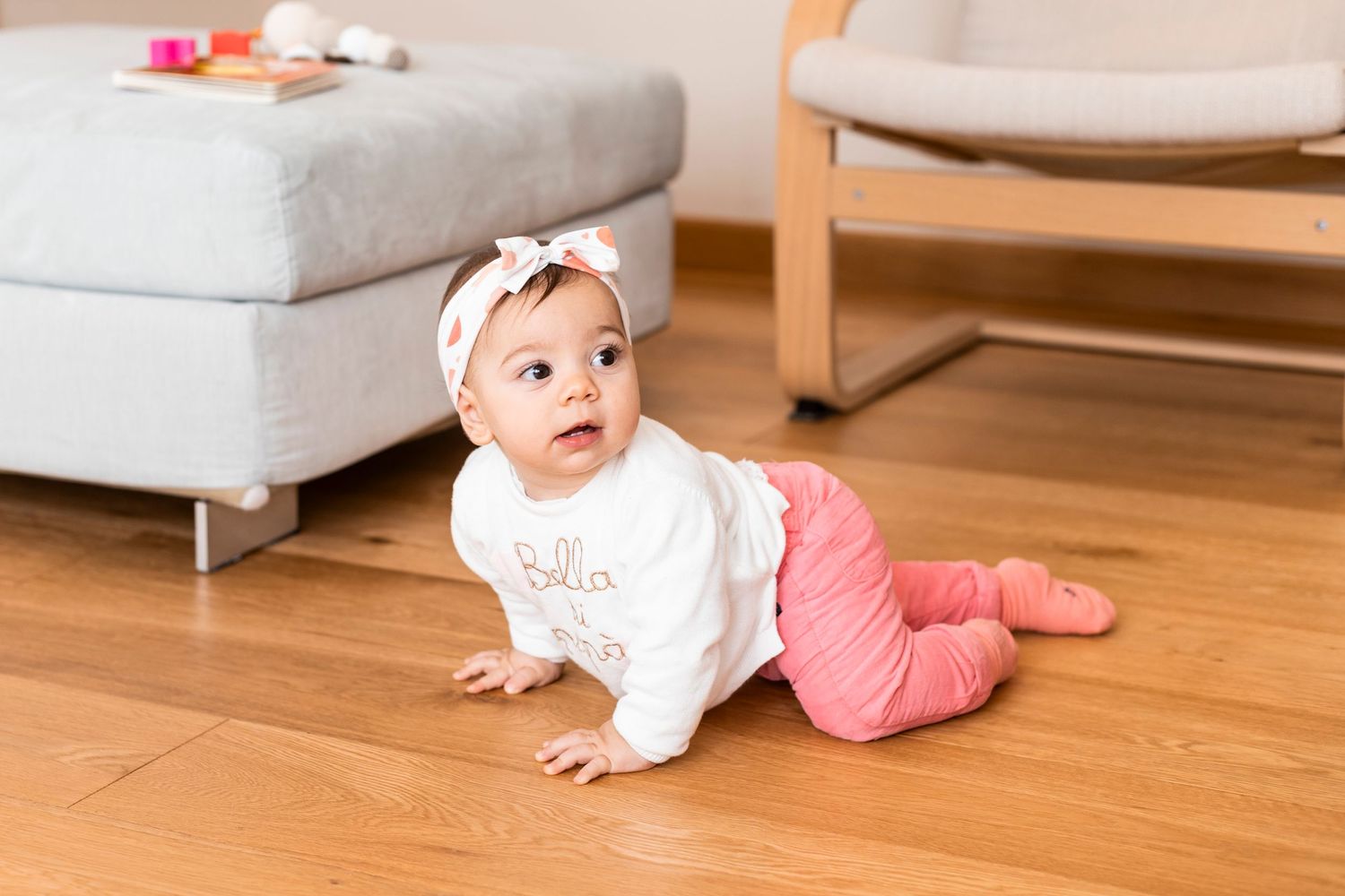Cute baby in pink outfit crawling on hardwood floor with headband bow.