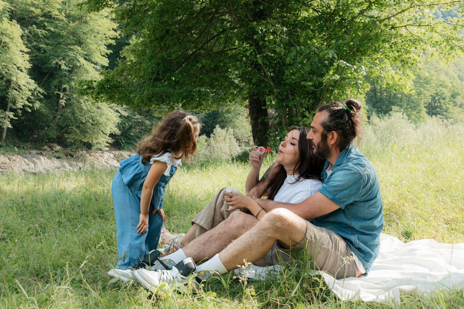 A happy family relaxes on a blanket in a green park, with their young daughter standing nearby.