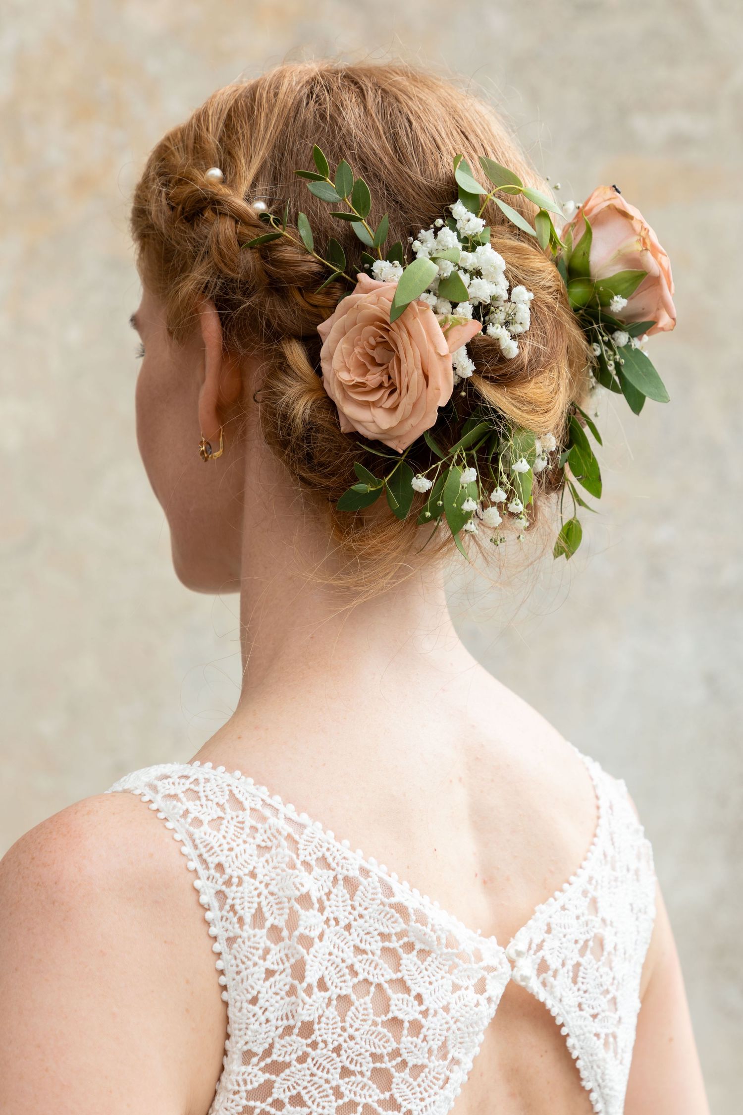 Bride with elegant updo hairstyle adorned with peach roses, baby's breath, and greenery, wearing white lace dress.