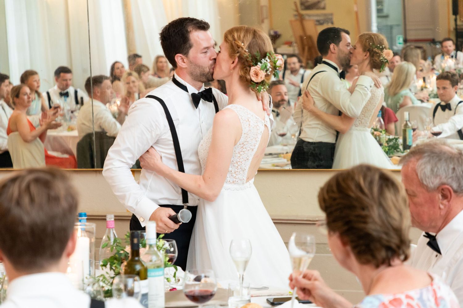 Bride and groom share romantic kiss during wedding reception while guests celebrate around them.