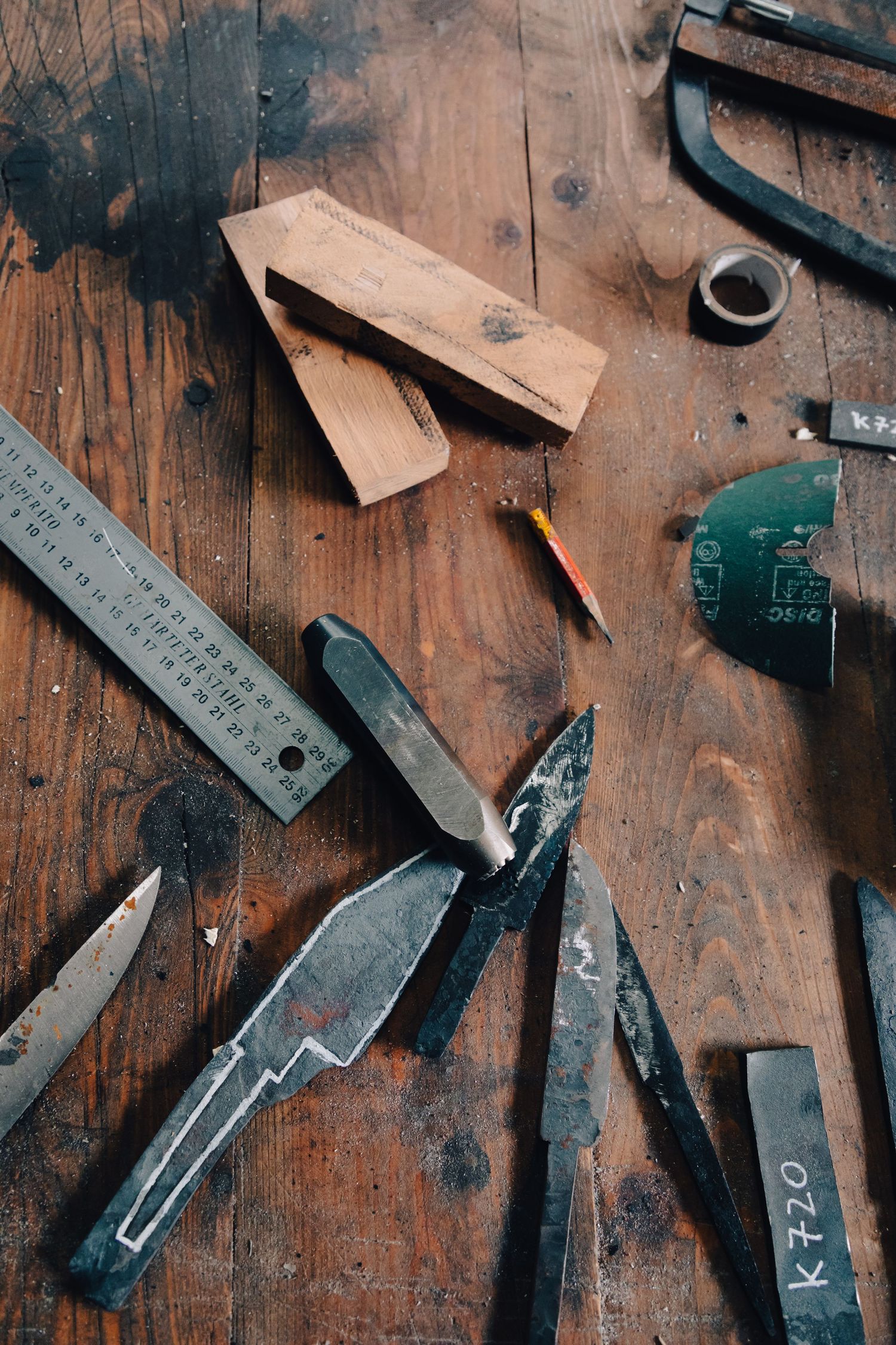 Flat lay of knife-making tools including blades, ruler, pencil, and sandpaper on wooden workbench.