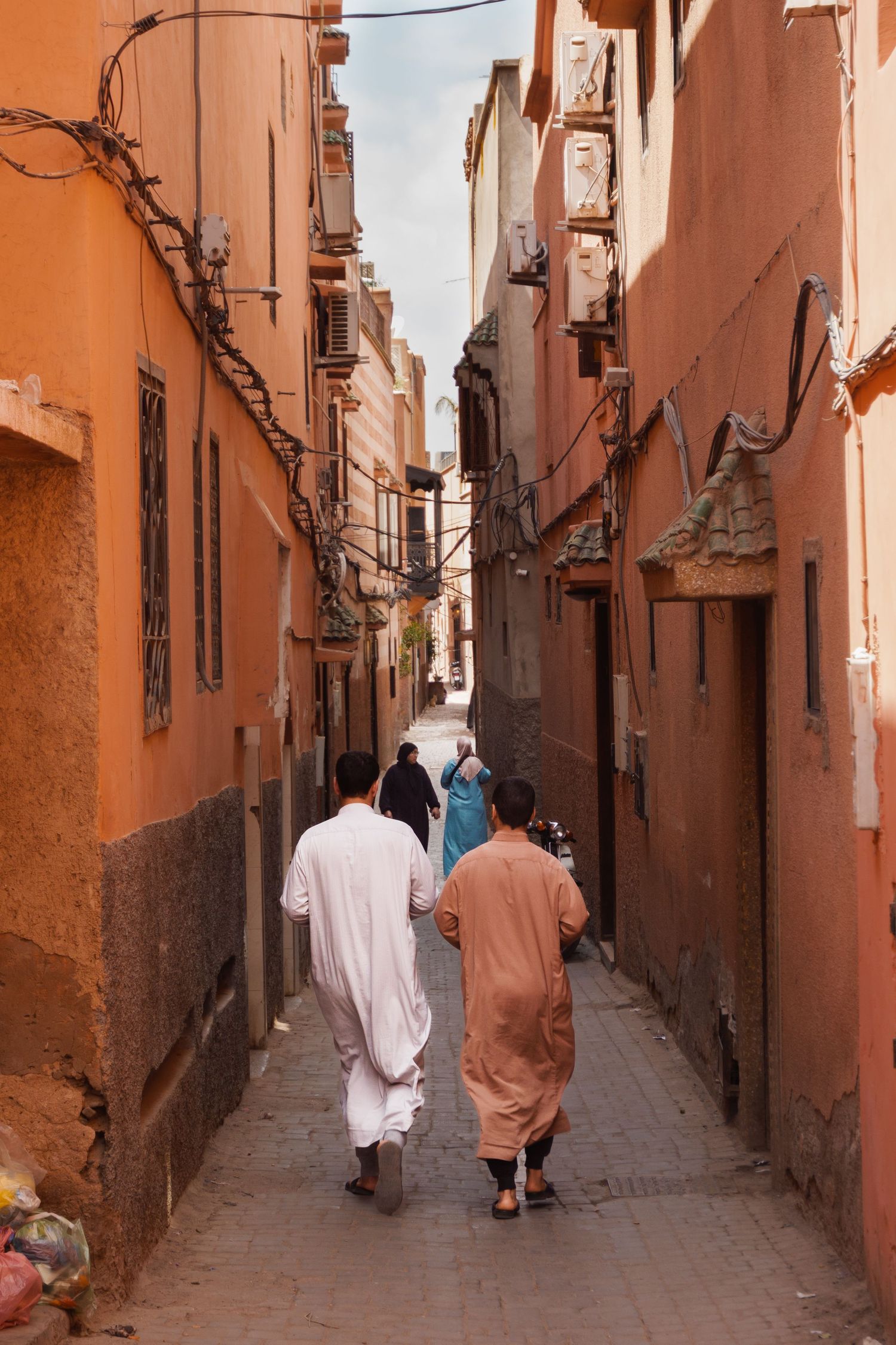 People in traditional djellabas walking through a narrow orange-walled alley in a Moroccan medina.