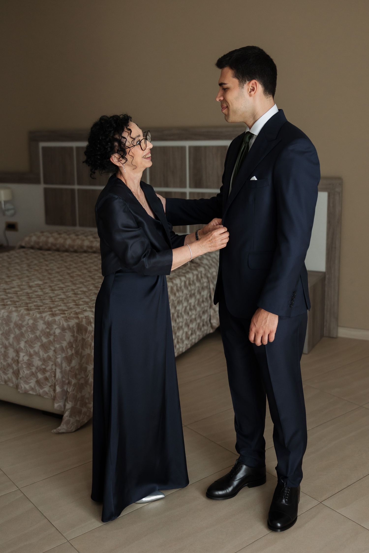 Older woman in black dress helping tall groom in navy suit adjust his boutonniere in a hotel room.
