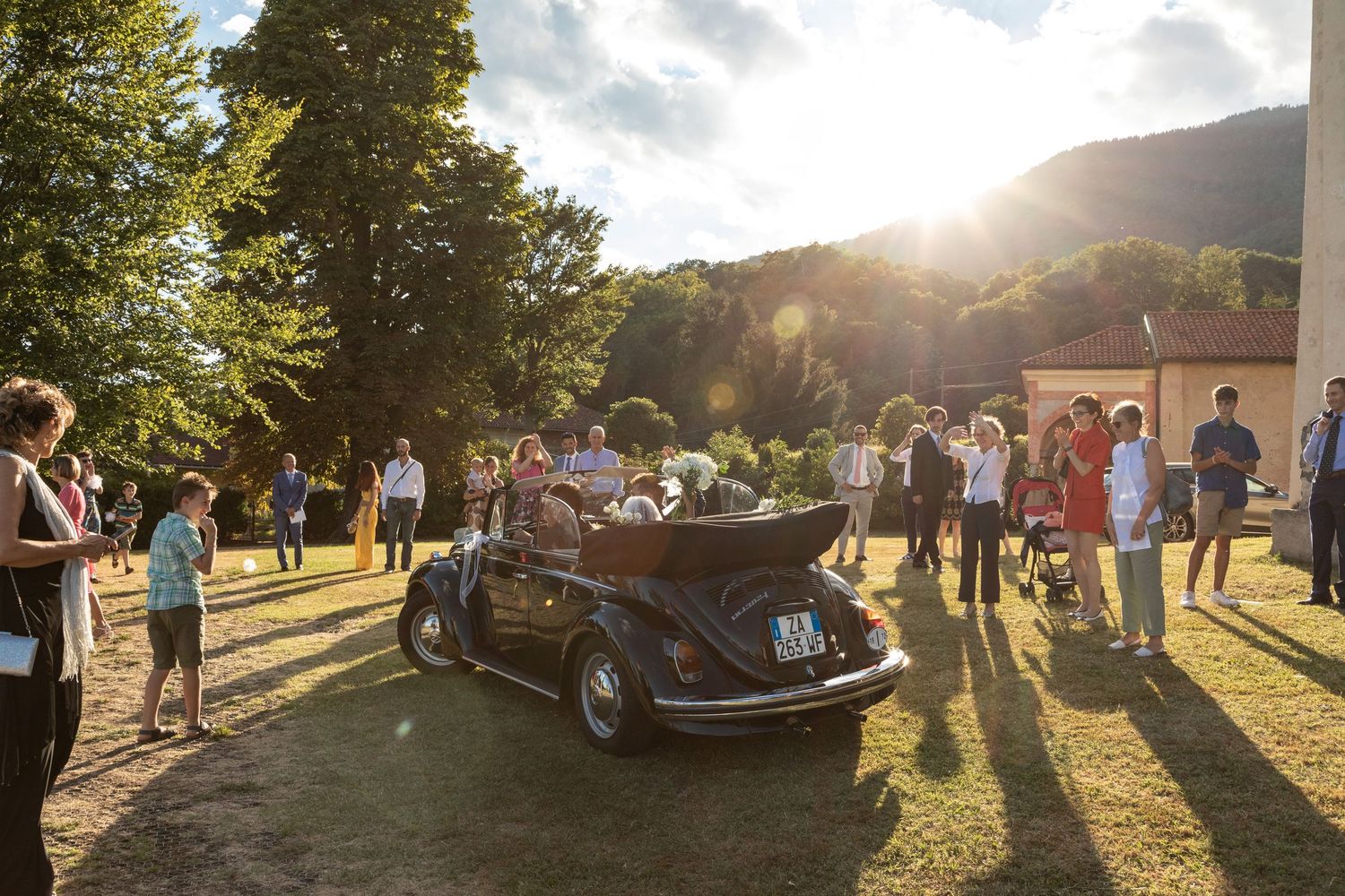 Wedding guests gather outdoors around a vintage black VW Beetle convertible in golden sunlight with mountains in background.