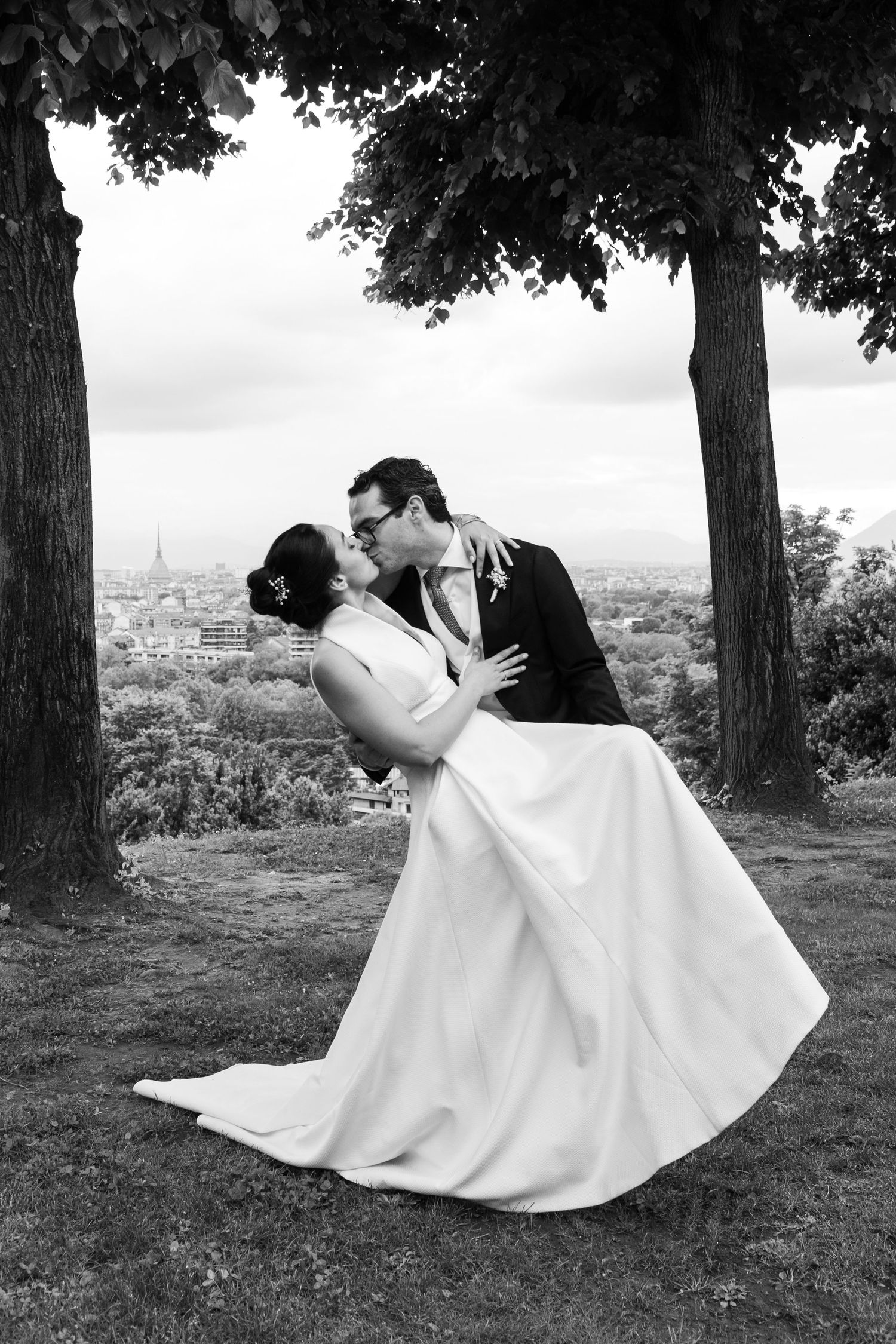 Black and white image of groom dipping bride for a kiss under trees with city panorama.