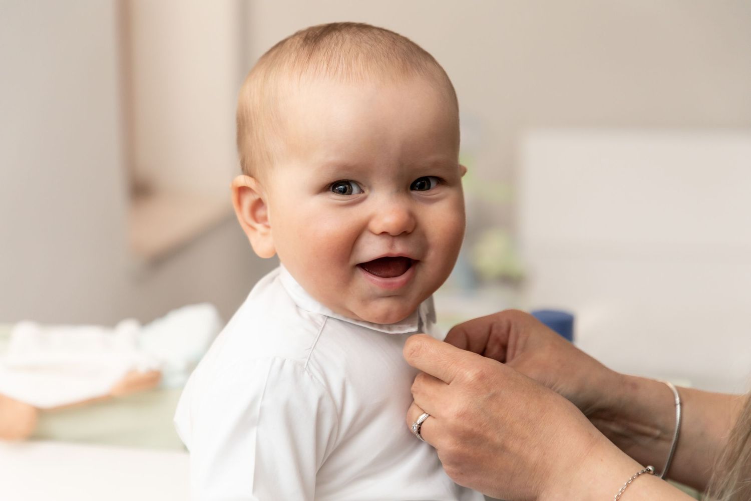 Smiling baby in a white outfit being held upright by adult hands in a bright room.