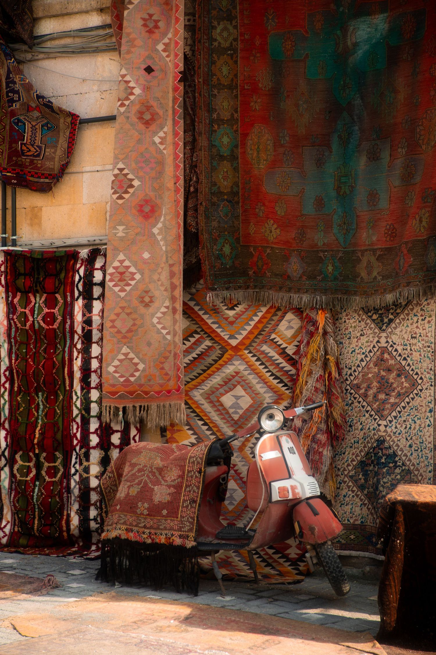 Colorful traditional Turkish kilim rugs and carpets displayed outside a market shop with a vintage red Vespa scooter.