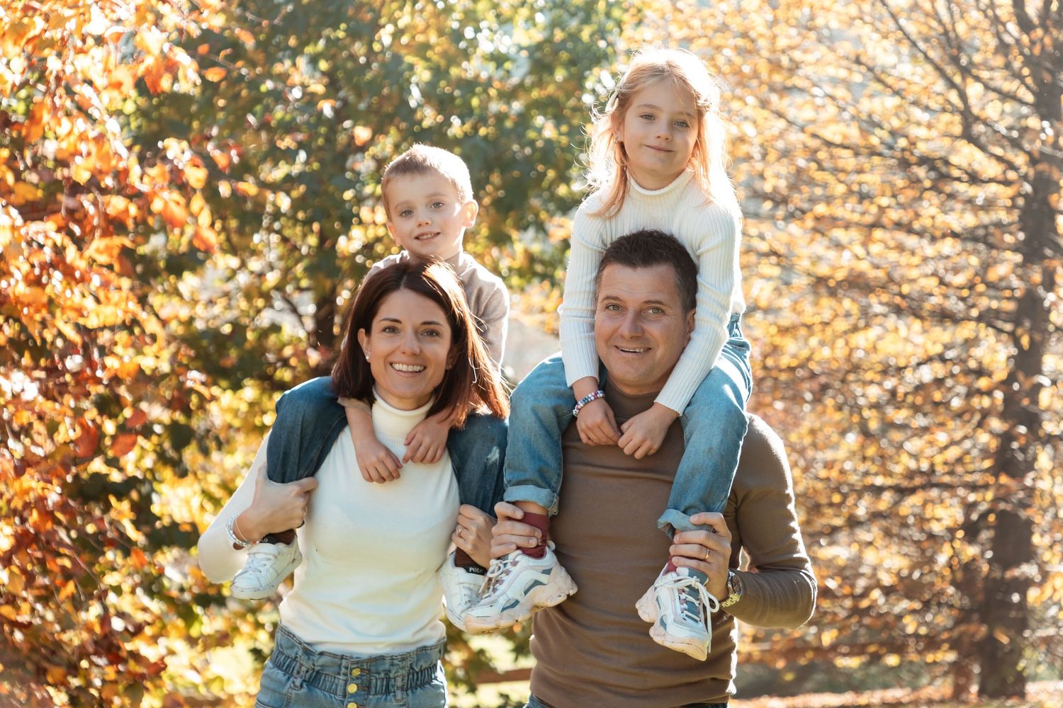A joyful family of four poses outdoors in autumn, with children riding on parents shoulders.