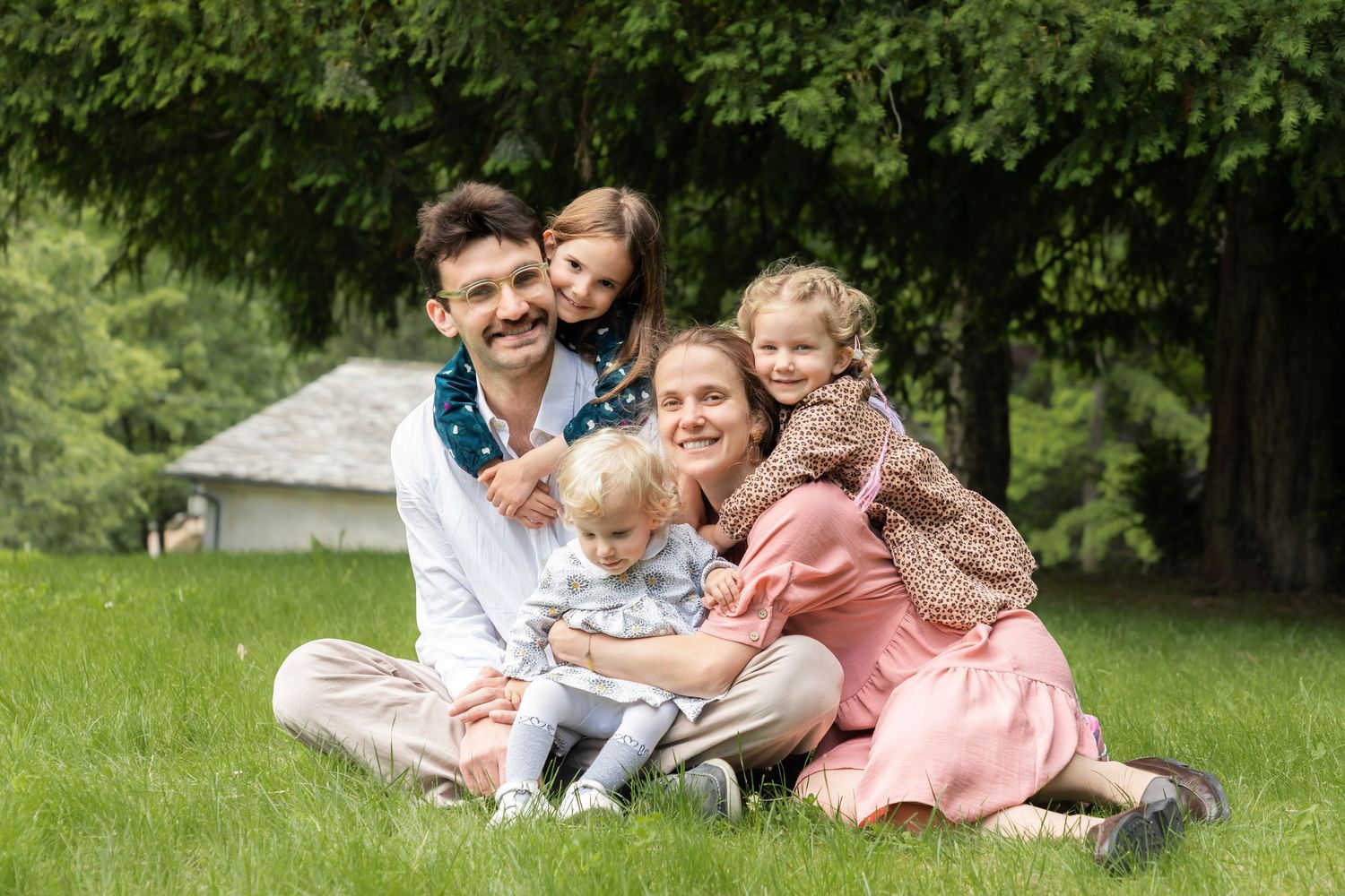 Joyful family of five sitting together on grass under green trees.