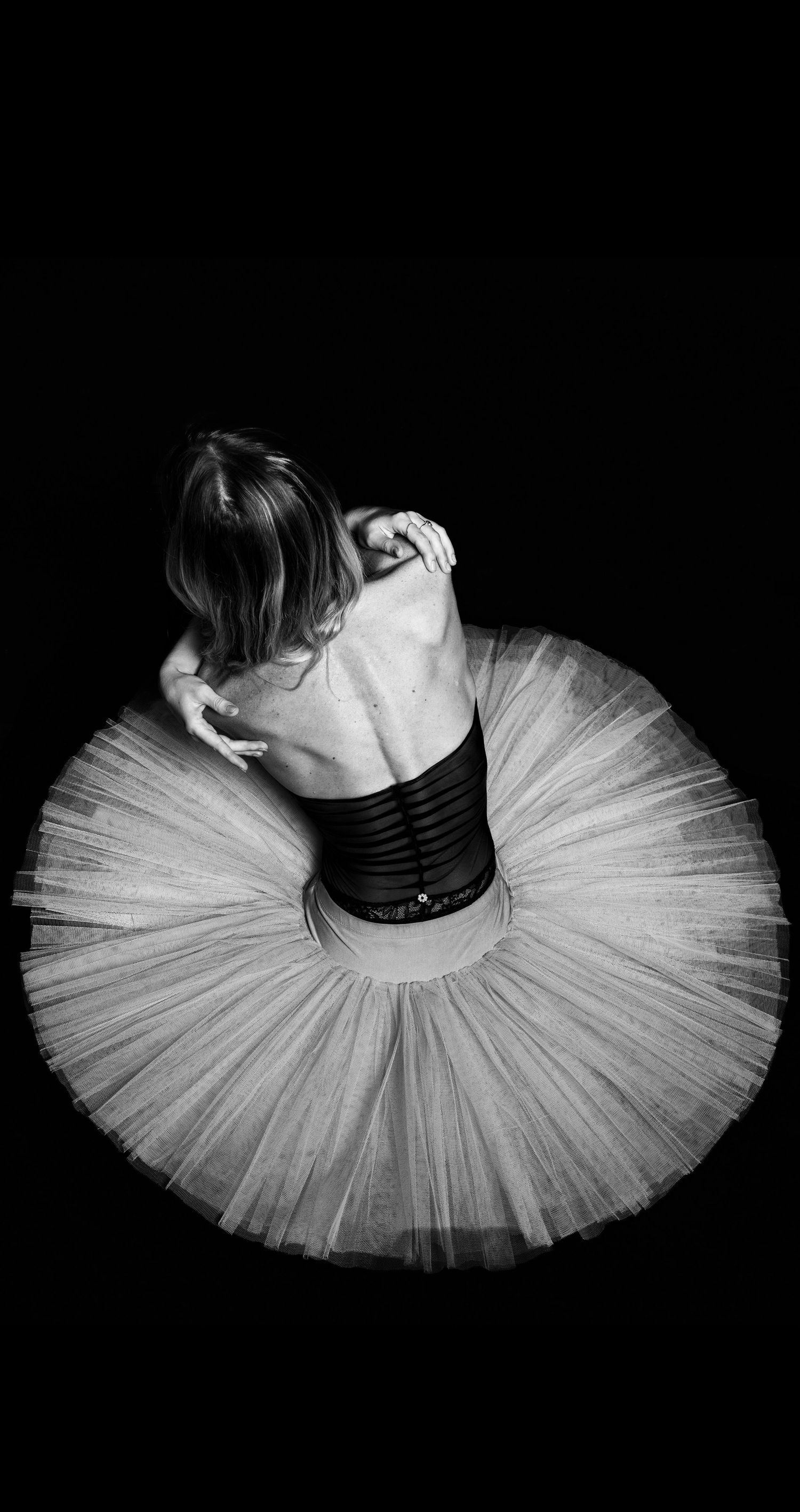 Overhead view of ballerina in white tutu hugging herself against dramatic black background.