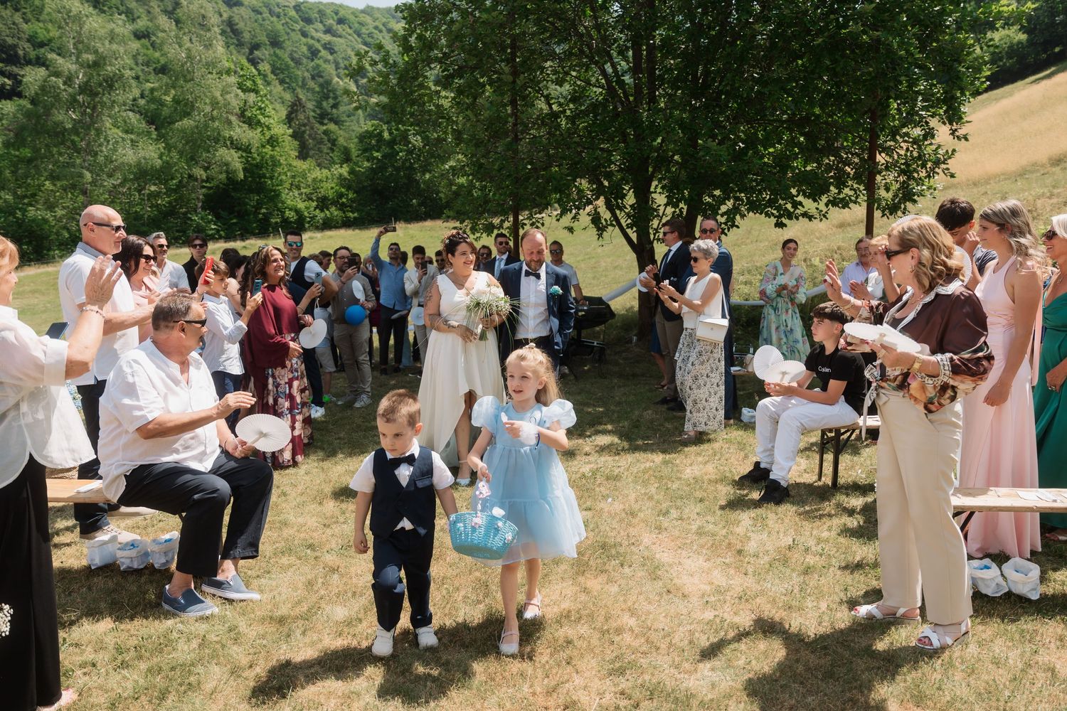 Flower girl and ring bearer walk down outdoor wedding aisle as guests celebrate in lush green countryside.