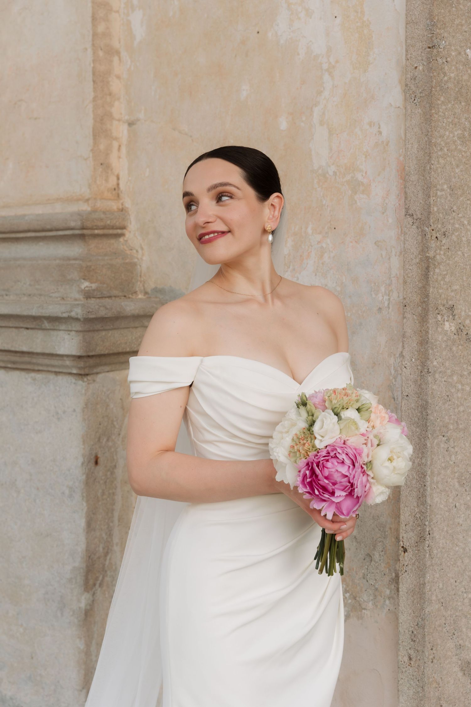 Elegant bride in off-shoulder gown holds pink and white peony bouquet against a stone wall.