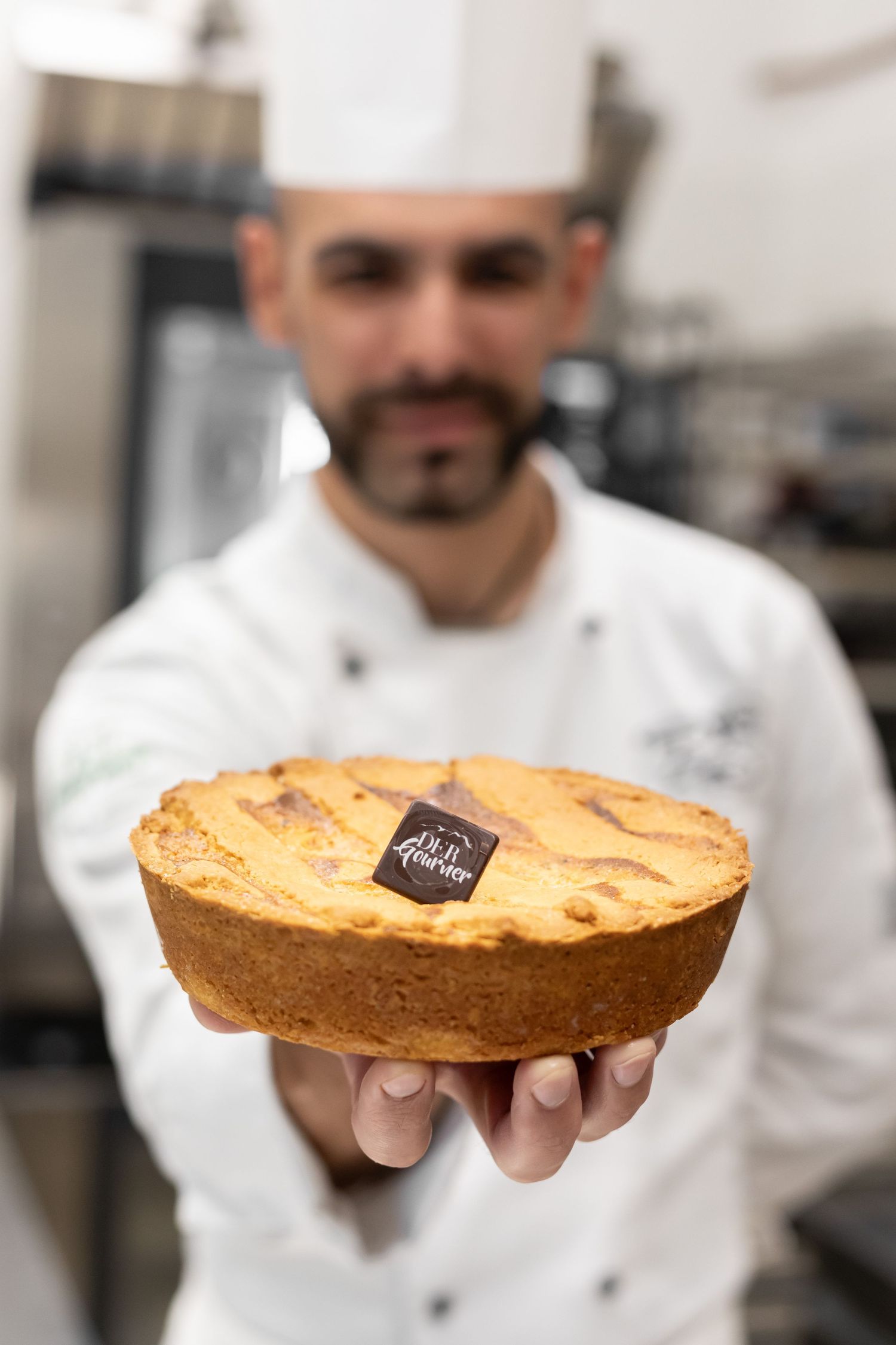 Pastry chef in white uniform and toque holding freshly baked golden tart with chocolate logo tag.